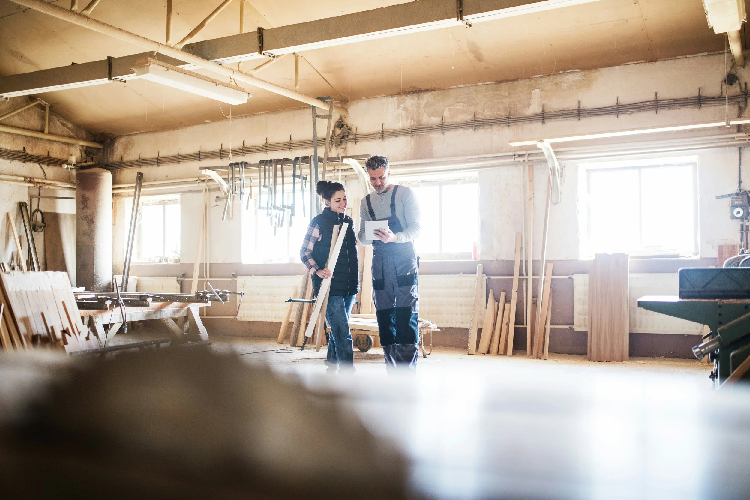 A man and a woman are standing inside a woodworking shop, looking at a tablet. They are surrounded by woodworking tools and wood pieces, with large windows letting in natural light.