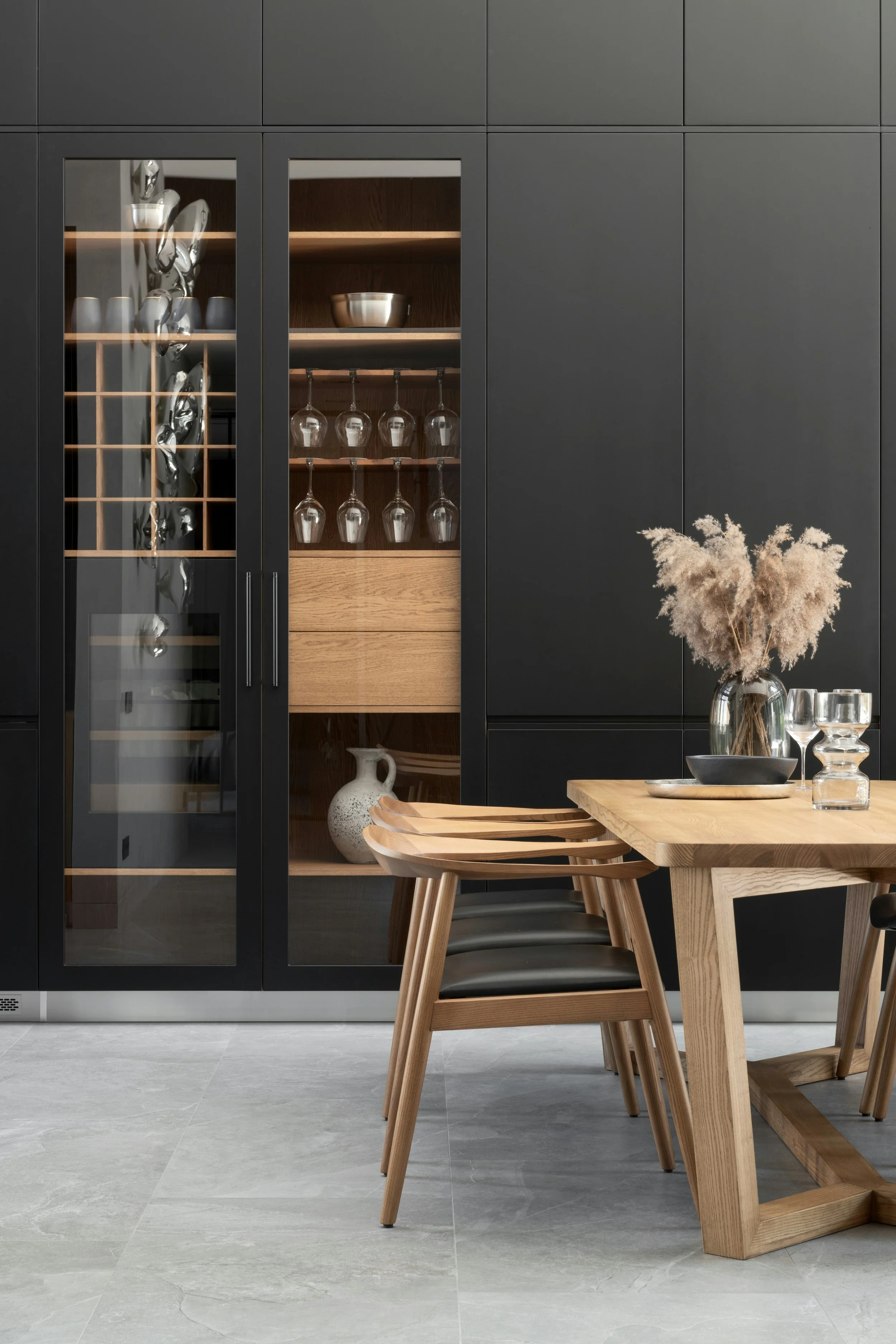A modern dining room with a wooden table and chairs, black cabinets with glass doors, and a vase with dried pampas grass on the table.