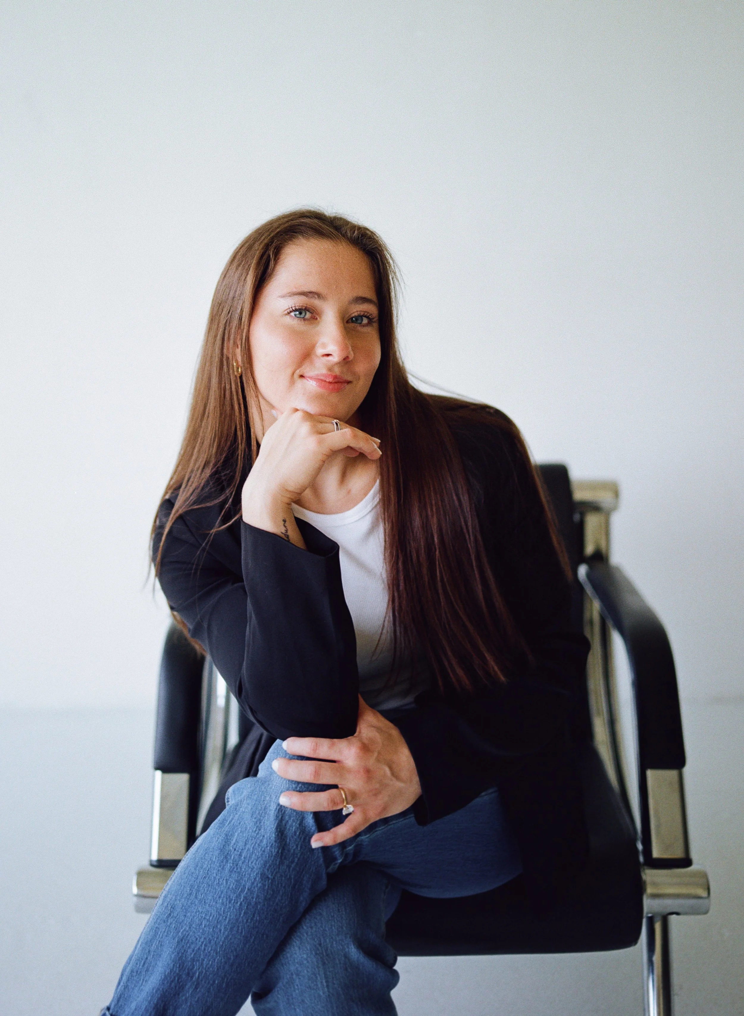 A woman with long brown hair, wearing a black blazer and white shirt, sitting on a black chair with her right leg crossed over her left, resting her chin on her hand and smiling slightly, against a plain light gray background.