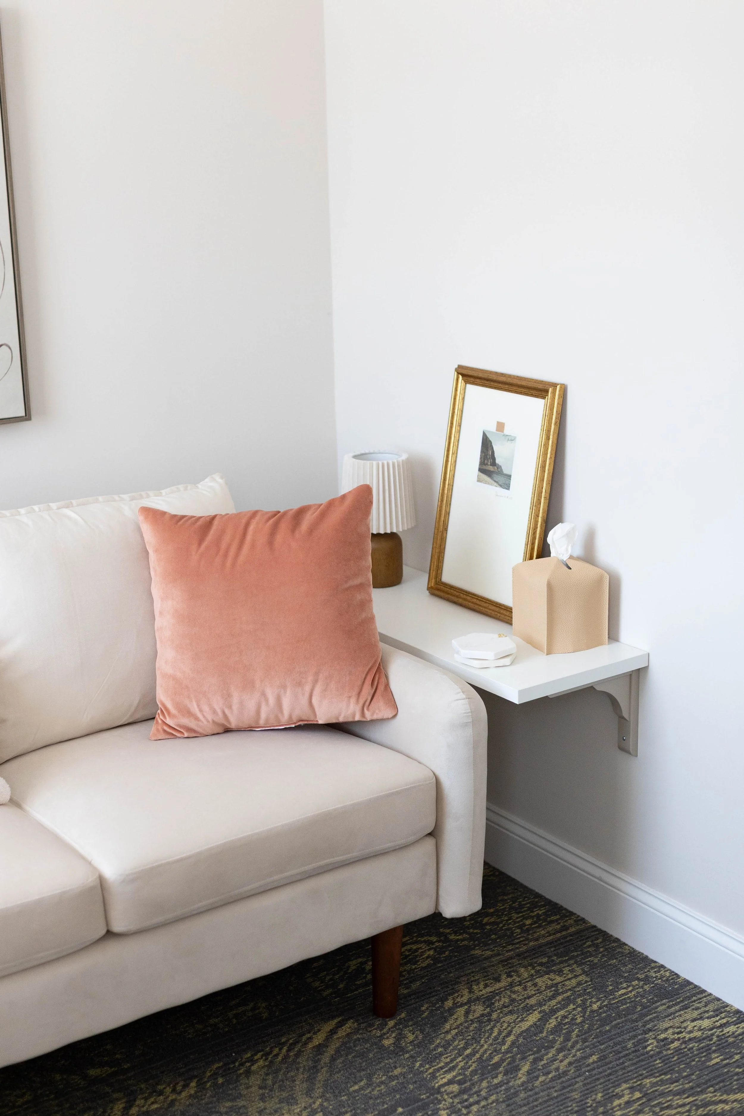 Corner of a cream loveseat with a coral velvet pillow beside a small wall-mounted shelf holding tissues and framed art, inside Shelley Snapp Therapy's Doylestown office