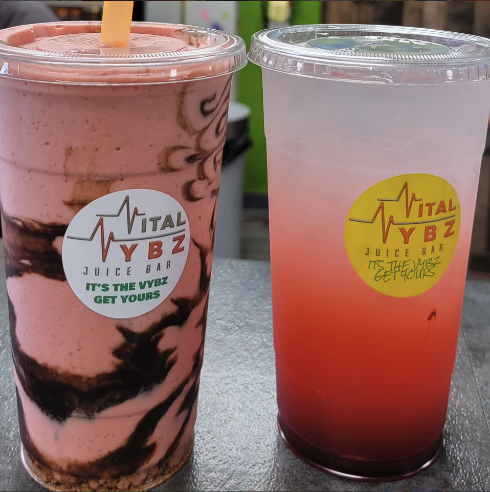 Two large colorful drinks in clear plastic cups with lids and straws from Vital YBZ Juice Bar. The left drink is pink with chocolate swirling, and the right drink is pinkish gradient with darker red at the bottom.