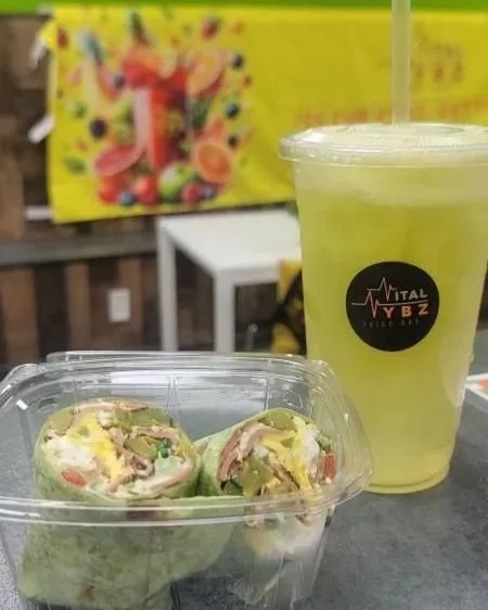 A clear plastic container with wraps or sandwiches and a large green beverage with a straw in a plastic cup, placed on a table with a colorful poster in the background.