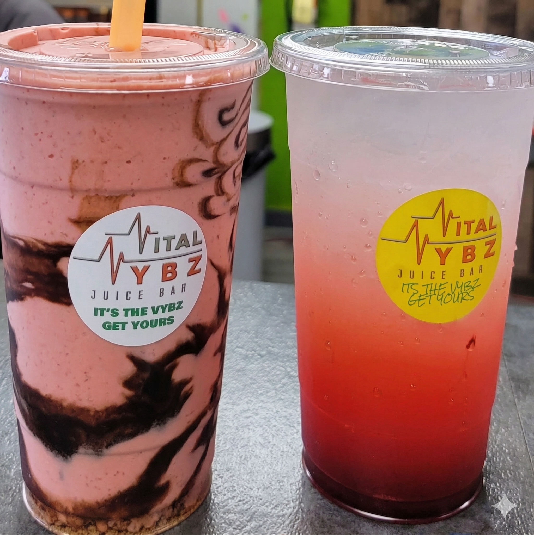 Two large plastic cups with seals, one containing a pink milkshake with chocolate syrup, and the other a pinkish-red drink with bubbles, both labeled with Vital YBZ Juice Bar logos.