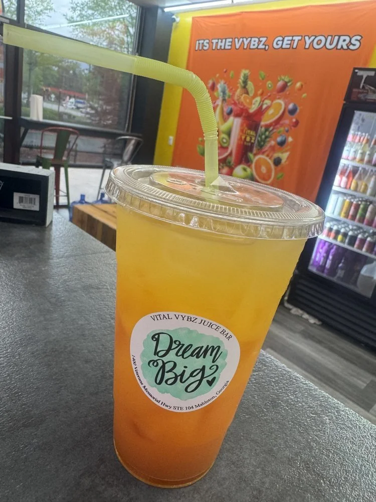 A large yellow-orange juice with a green straw in a clear plastic cup, labeled 'Dream Big' from Vital Vybz Juice Bar, with a colorful mural and fridge stocked with drinks in the background.