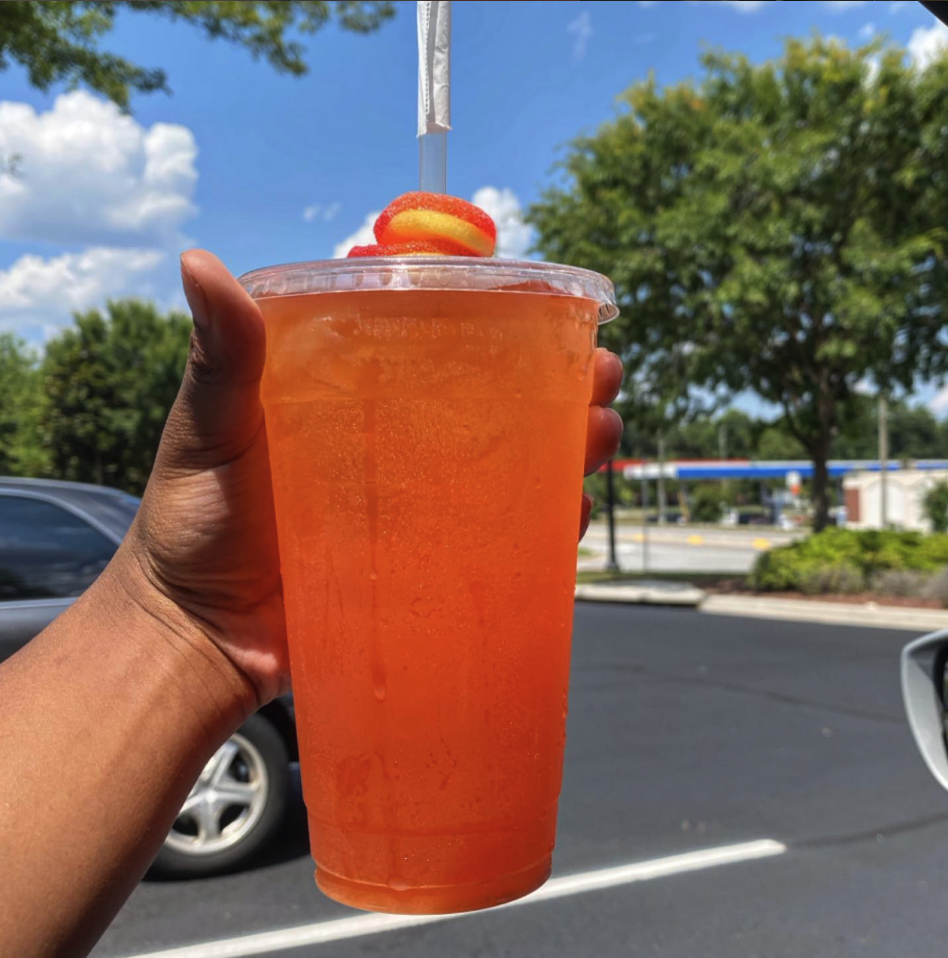 Close-up of a hand holding a large clear plastic cup filled with an orange-colored beverage, topped with a swirl of red and yellow candy and a straw, against a background of a parking lot, trees, a blue sky with some clouds.