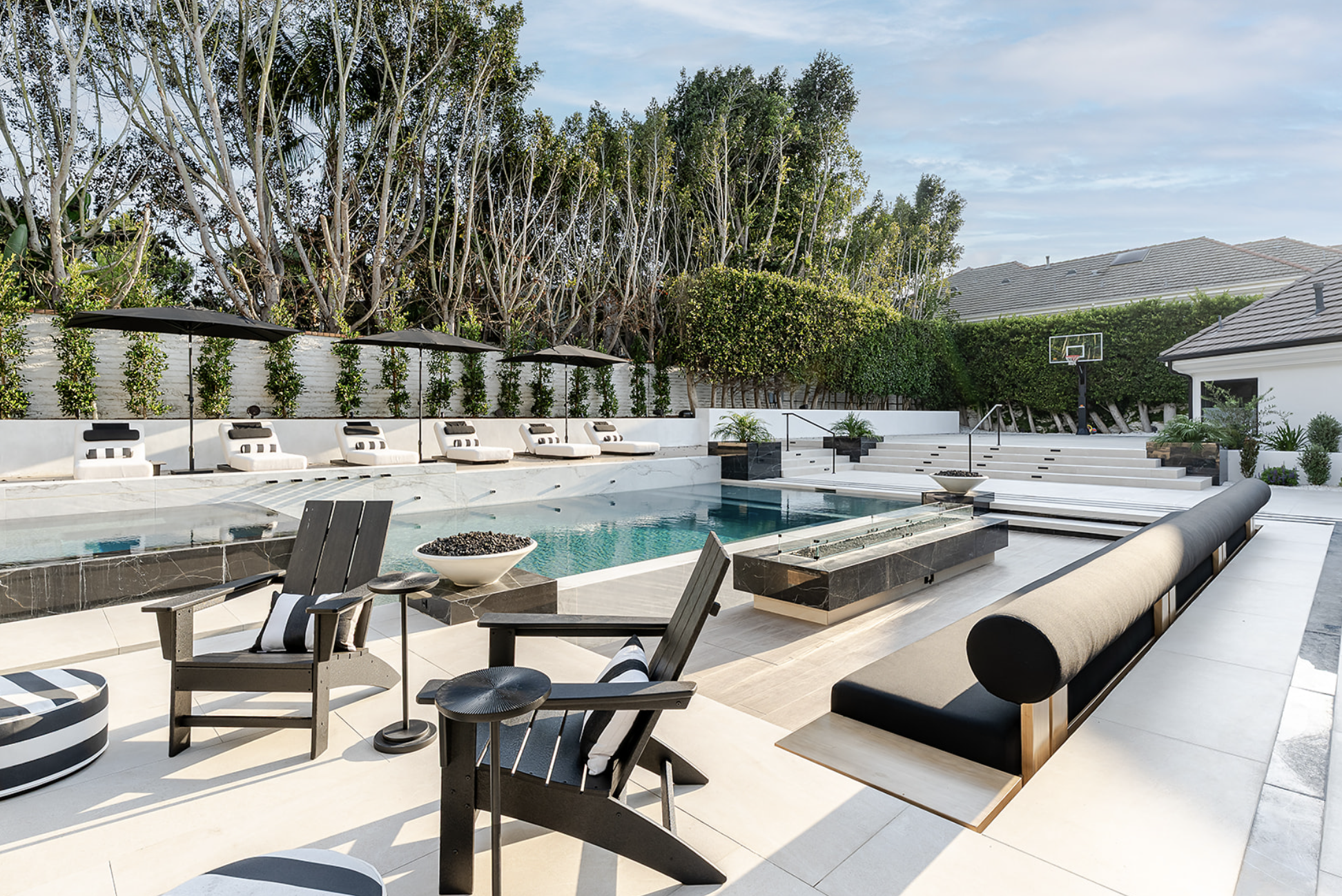 Modern backyard with a swimming pool, lounge chairs with umbrellas, trees, and a basketball hoop.