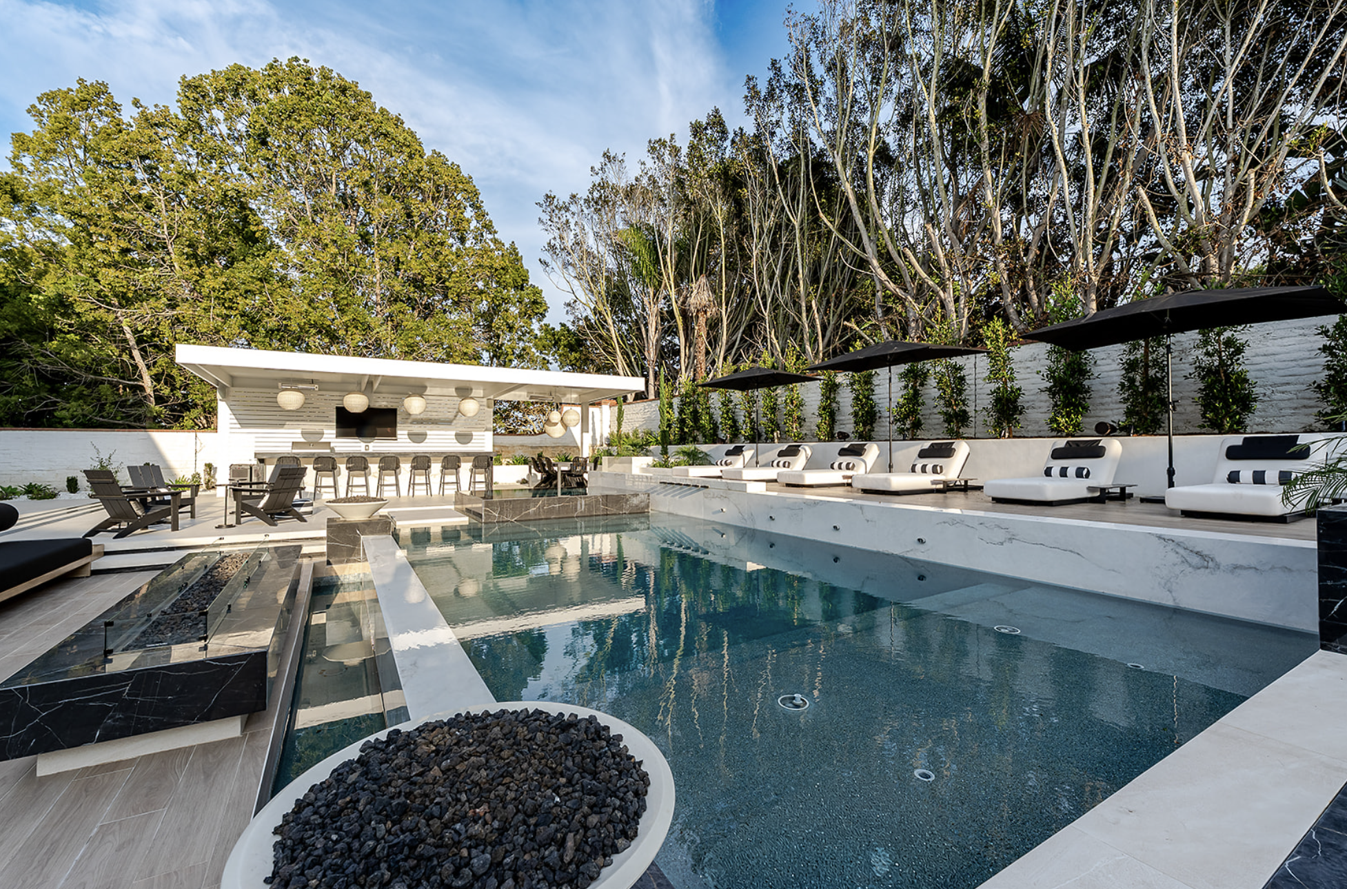 Outdoor pool area with white lounge chairs under black umbrellas along a high white wall with green plants, surrounded by large trees and a partly cloudy sky.