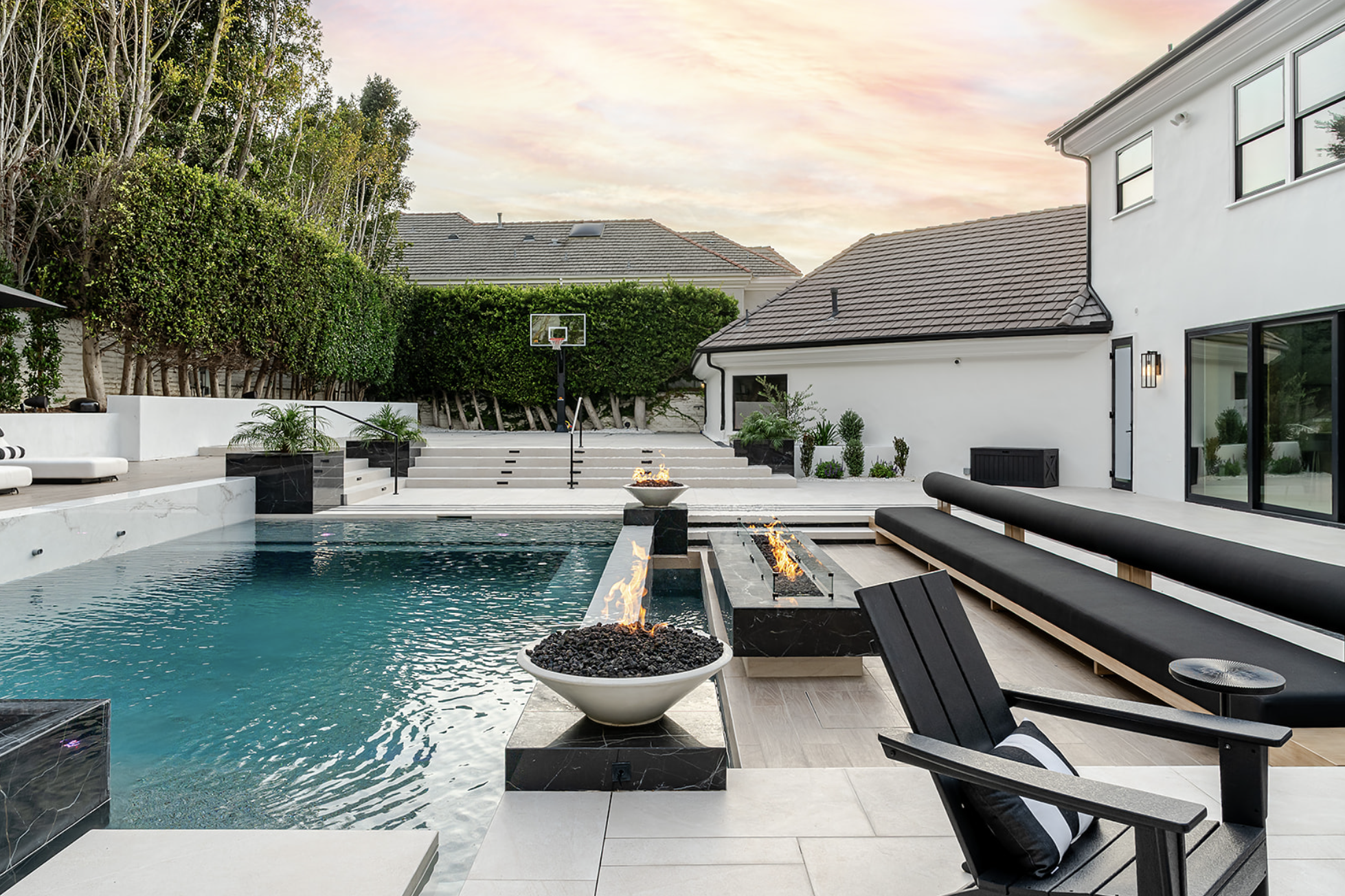 Modern backyard with a swimming pool, fire pit, and patio furniture. Features luxury lounge chairs, small black tables, and a fire bowl. Lush green trees and residential rooftops are in the background, with a pastel sky at sunset.