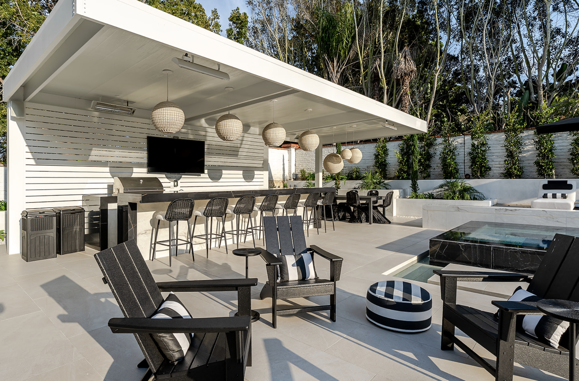 Outdoor patio with black chairs, a bar counter with high chairs, hanging paper lantern lights, a large flat-screen TV, a built-in grill, lush green trees and plants in the background, and a seating area near water features.