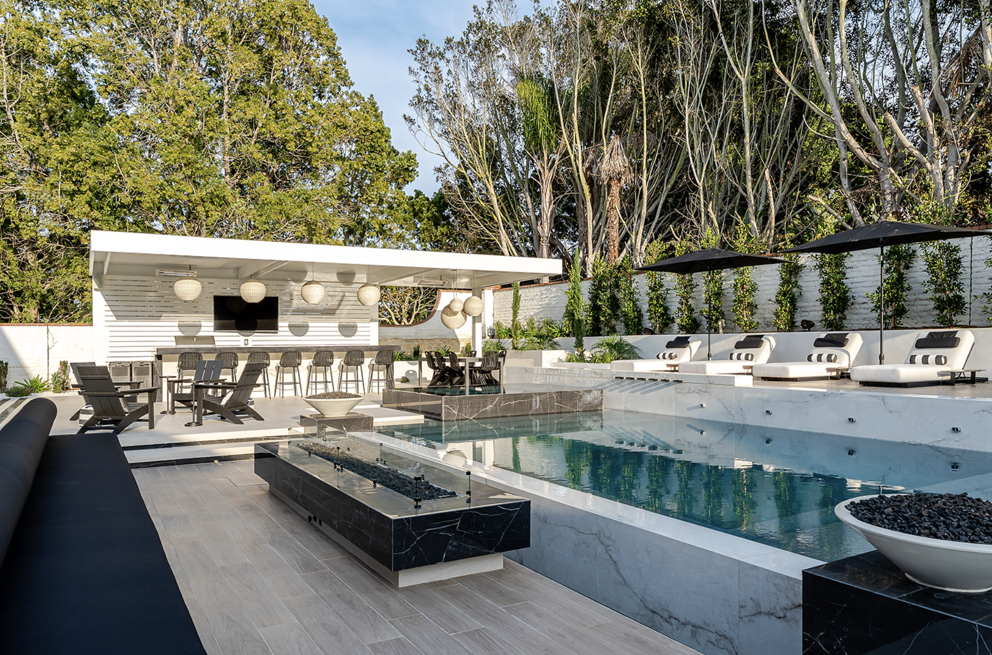 Modern backyard with outdoor seating, a pool, and a white privacy wall with lush trees behind.