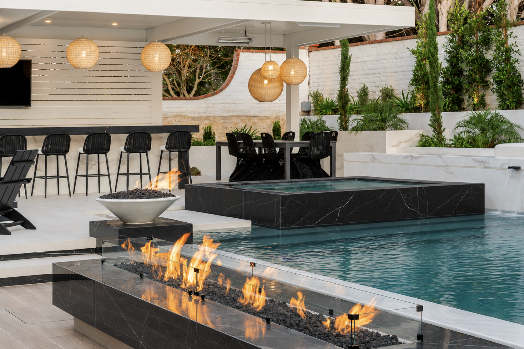A modern outdoor patio with a swimming pool, a fire feature, black bar stools, a dining table with chairs, hanging wicker pendant lights, lush green plants, and a white brick wall.