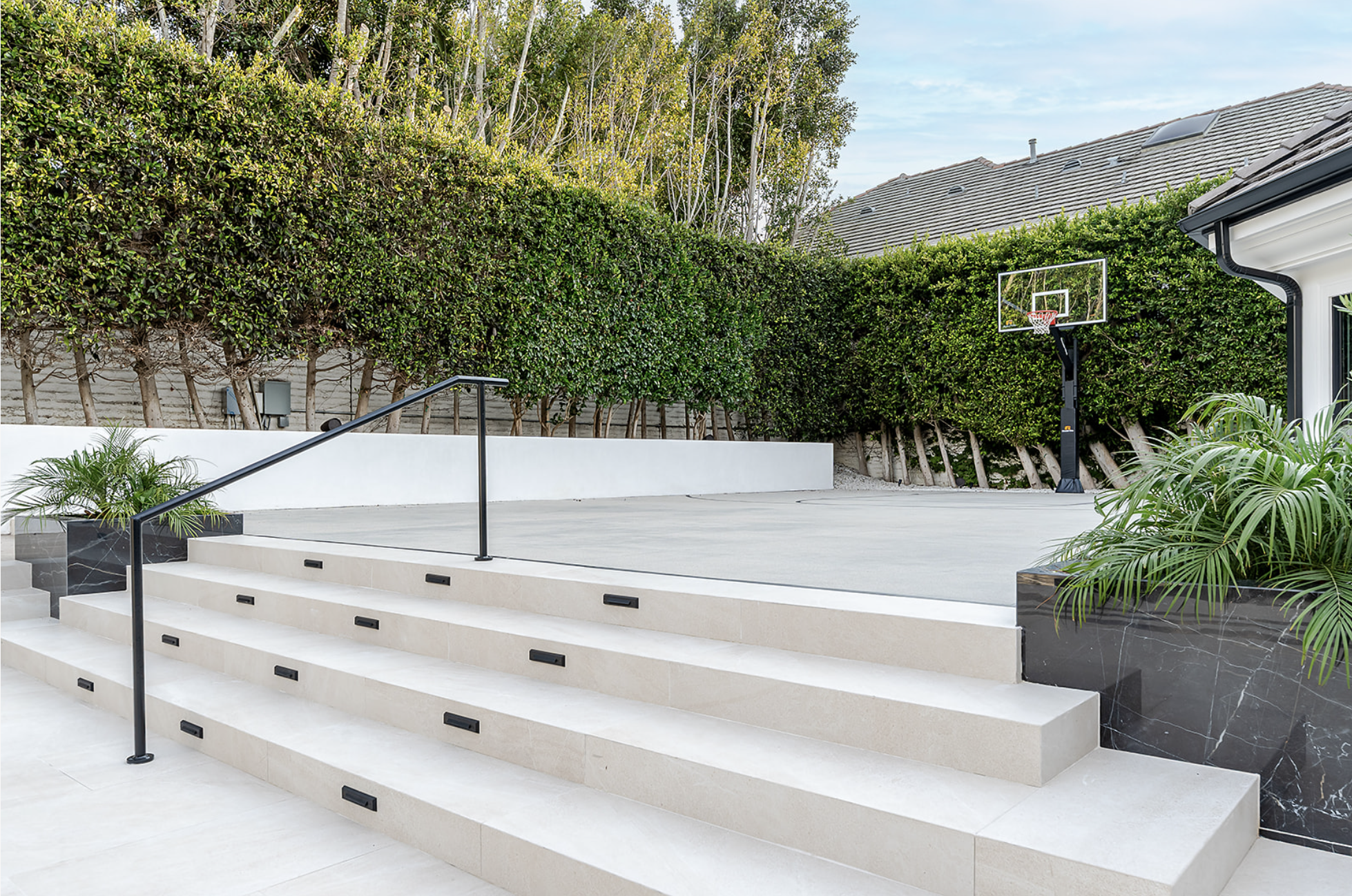 Modern outdoor basketball court with steps, black metal railing, landscaped plants, tall green hedge, house with tiled roof, clear blue sky