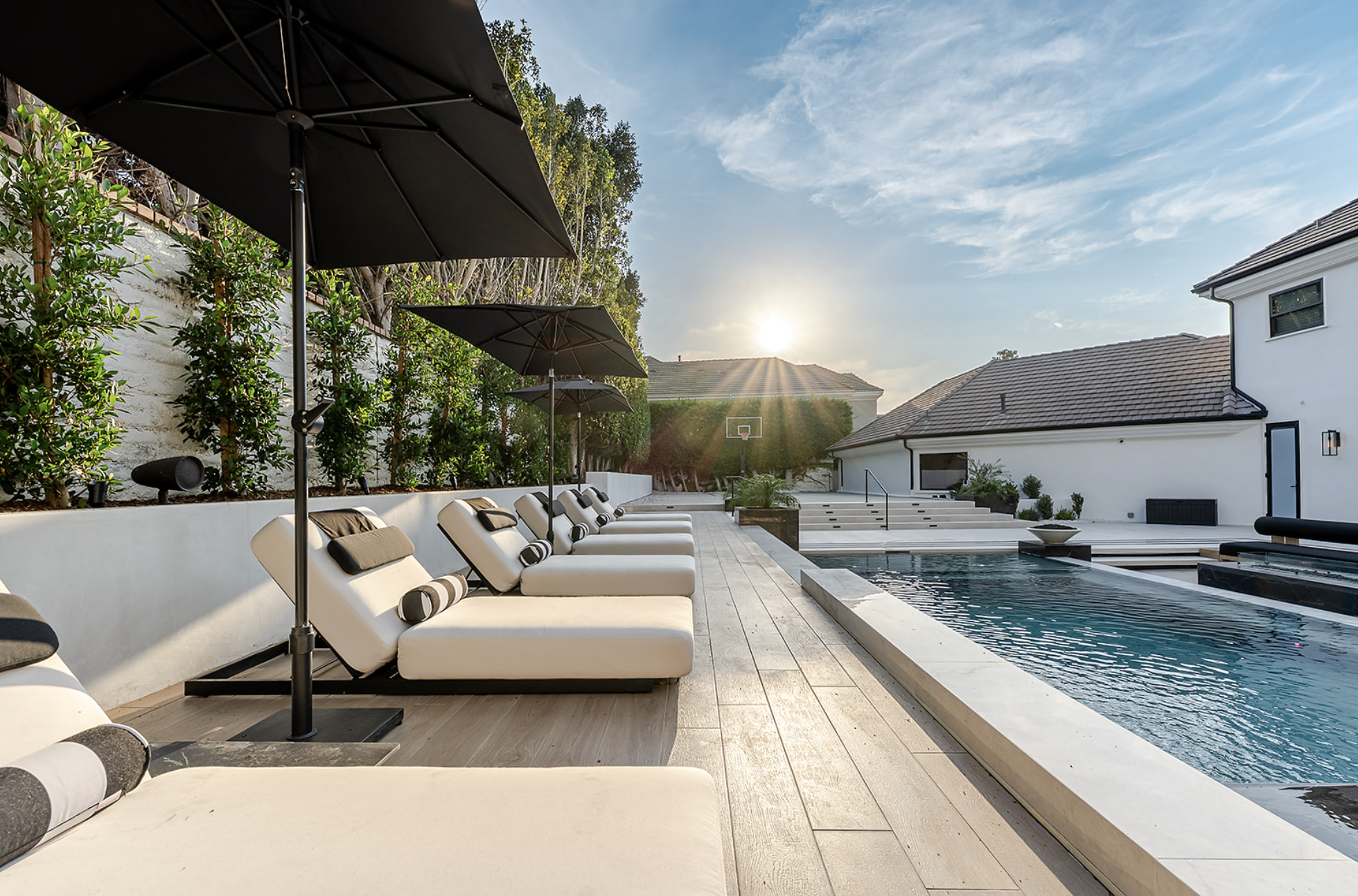 A luxurious backyard pool area with white lounge chairs, black umbrellas, and a modern white house with a sloped roof. The pool is rectangular with steps leading into it, and there is a basketball hoop in the background. The scene is set during sunset with a clear sky and sunshine.