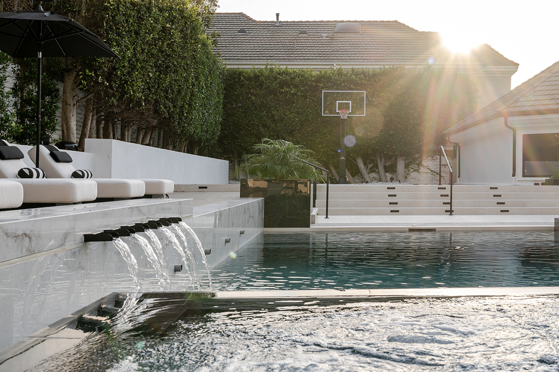 A backyard swimming pool with a waterfall feature, white lounge chairs with black cushions and a large black umbrella, surrounded by greenery and a basketball hoop in the background, with sunlight brightening the scene.