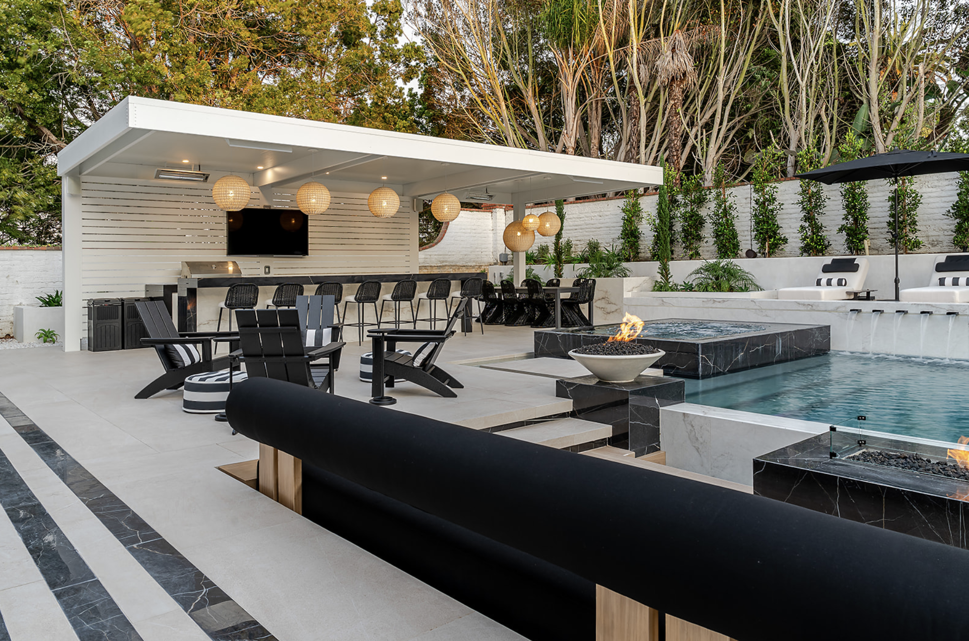 Modern backyard patio with pool, fire feature, black outdoor furniture, bar area, and string pendant lights, surrounded by greenery and trees.