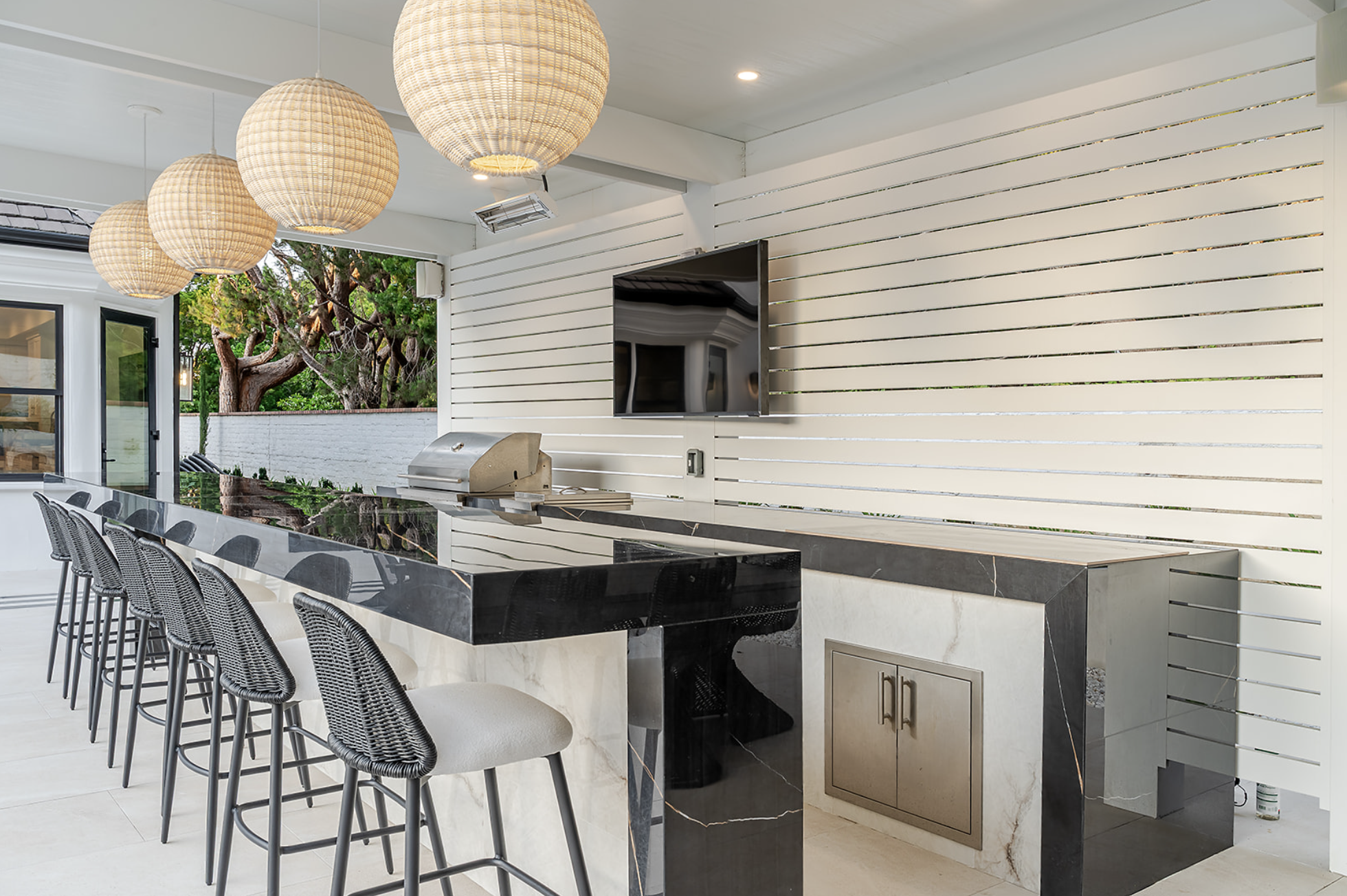 Outdoor kitchen area featuring a large black granite dining table with woven chairs, a built-in grill, and a white slatted wall with a mounted flat-screen TV. Decorative wicker pendant lights hang from the ceiling, and there is a view of green trees outside.