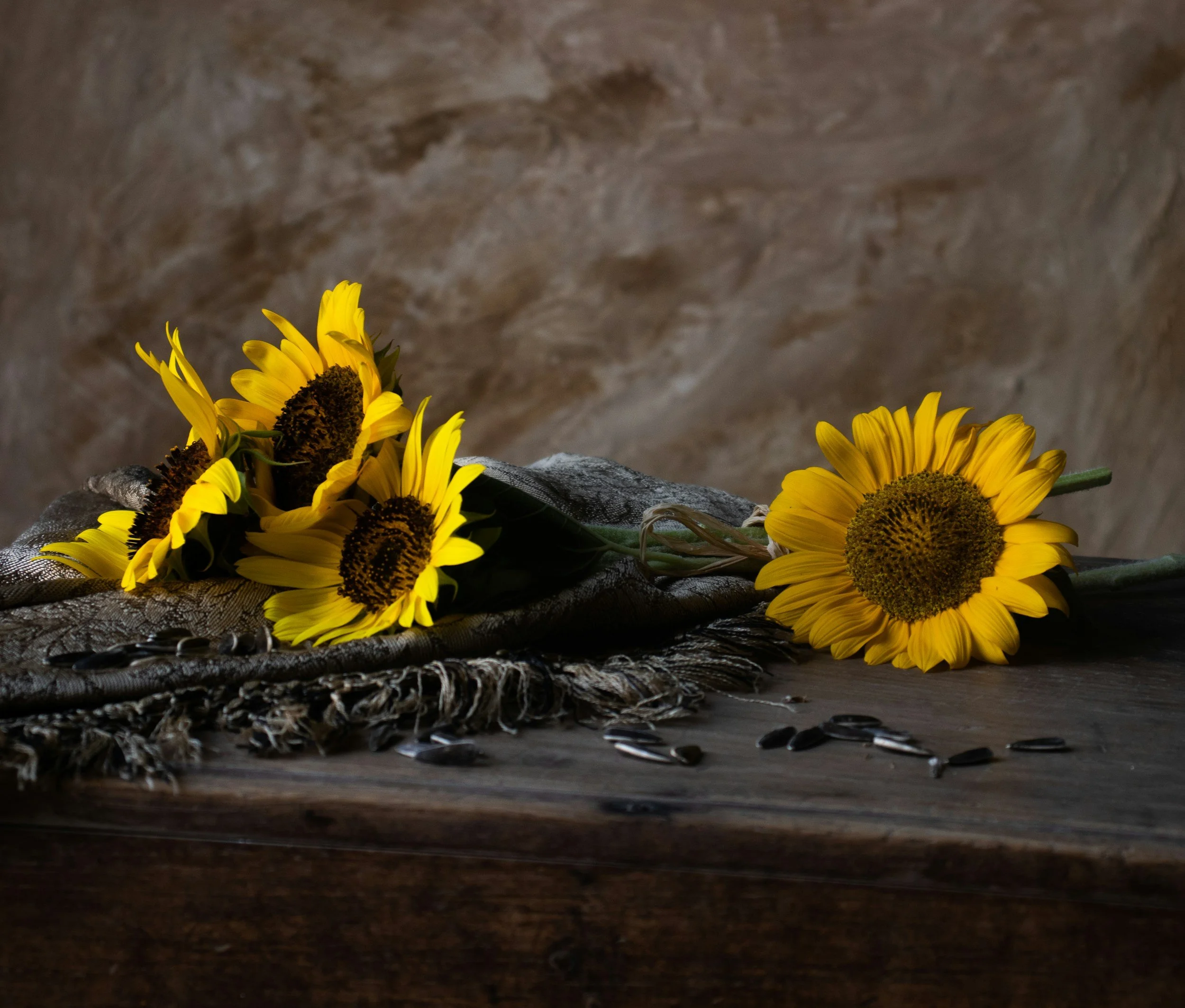 A bouquet of yellow sunflowers placed on a dark wooden surface, with sunflower seeds scattered around, against a textured brown background.