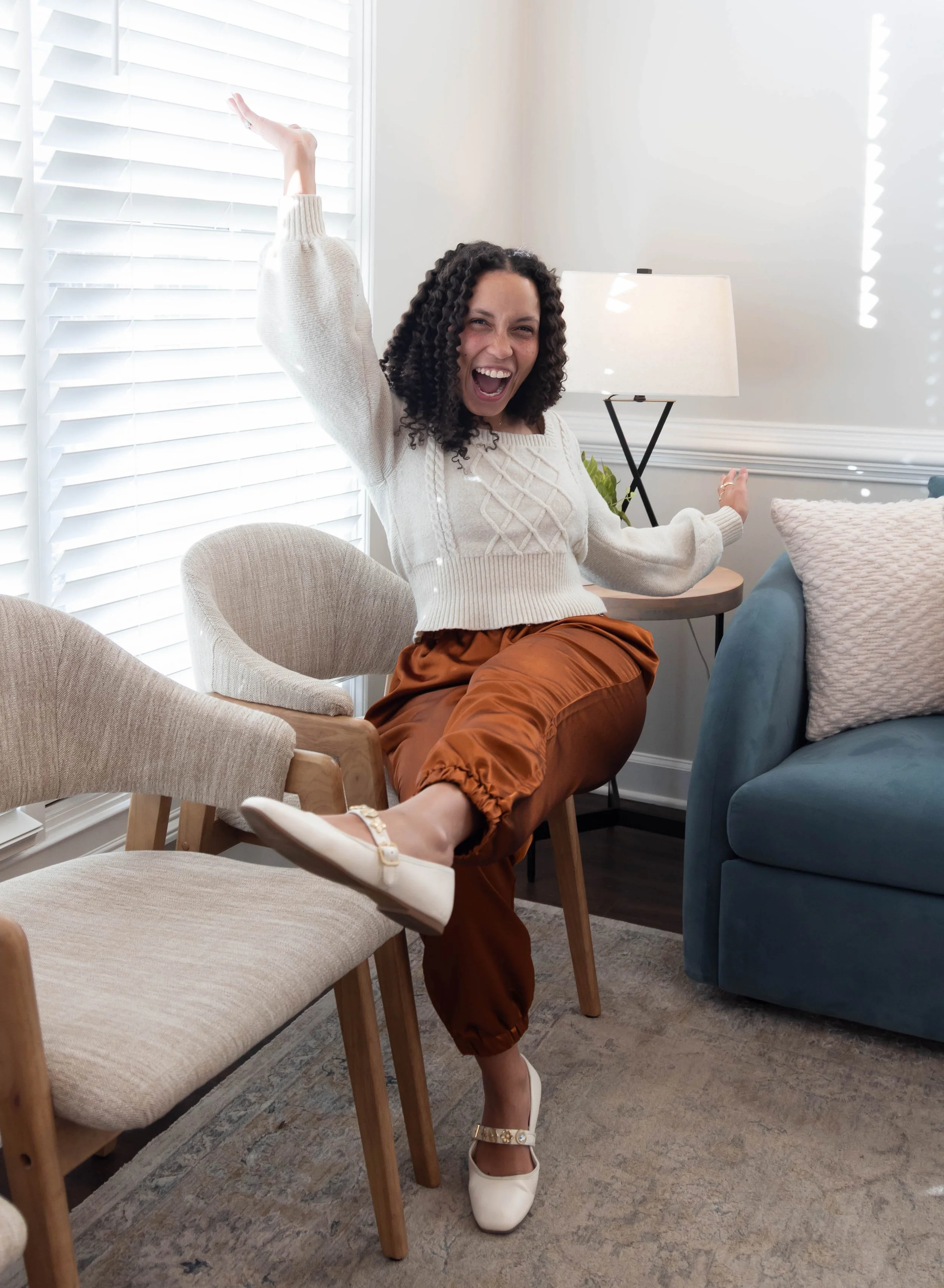 A woman with curly hair and a big smile, sitting on a chair with one leg up on another chair, in a bright living room, gesturing enthusiastically.