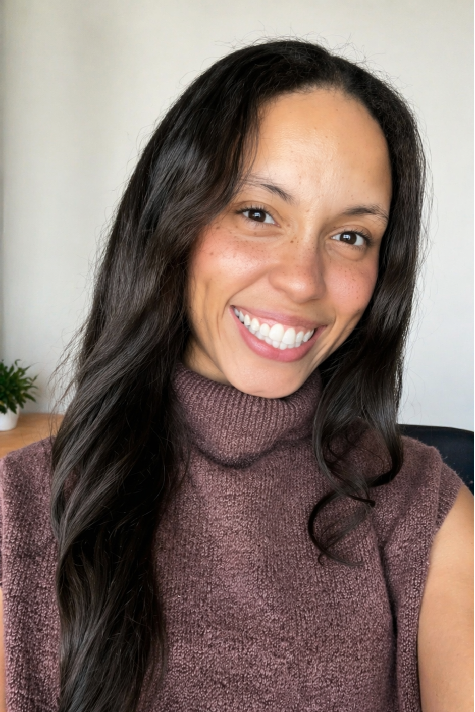 Close-up photo of a smiling woman with long dark hair, wearing a purple turtleneck sweater, in a bright room with a small potted plant in the background.