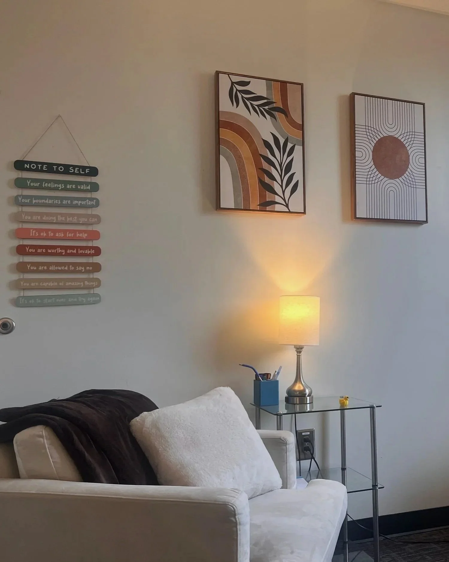 A cozy living room corner featuring a cream-colored sofa with a black and a white pillow, a glass side table holding a silver lamp, a blue tissue box, and a yellow toy. The wall has two framed abstract art pieces and a decorative note to self with motivational messages.