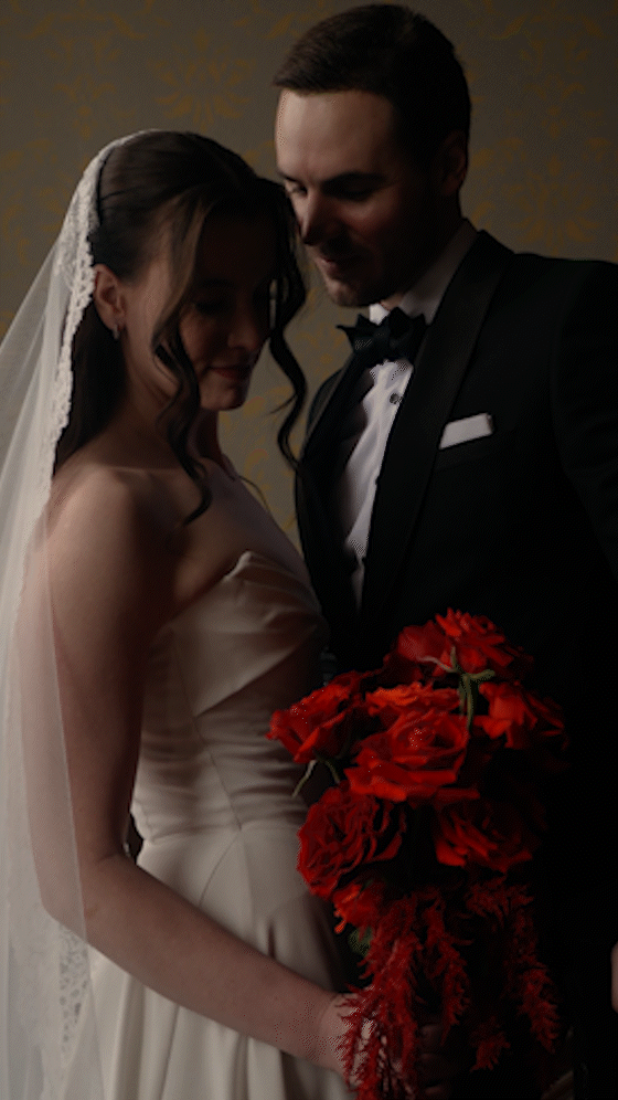 A bride and groom on their wedding day, standing close together and sharing a tender moment. The bride is wearing a white wedding gown and veil, holding a bouquet of red flowers. The groom is in a black tuxedo with a bow tie.