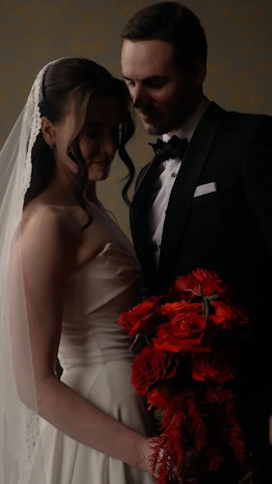A bride and groom on their wedding day, standing close together and sharing a tender moment. The bride is wearing a white wedding gown and veil, holding a bouquet of red flowers. The groom is in a black tuxedo with a bow tie.