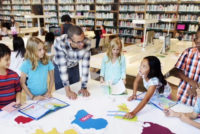 Children with an adult gathered around a table in a library, looking at maps and books.