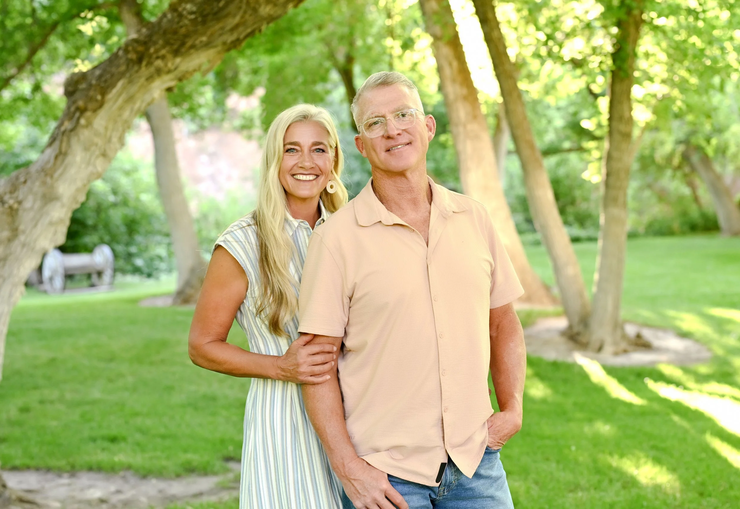 A smiling woman with long blonde hair and earrings stands behind a man with glasses, short blonde hair, and a light peach shirt, in a park with green trees and grass.