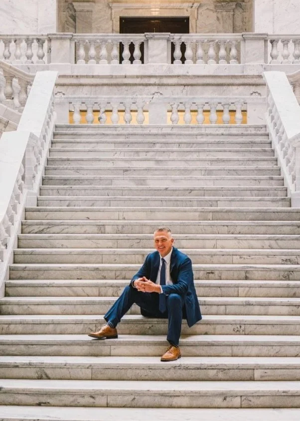 A man in a navy suit and brown shoes sitting on marble stairs, smiling while holding a phone.