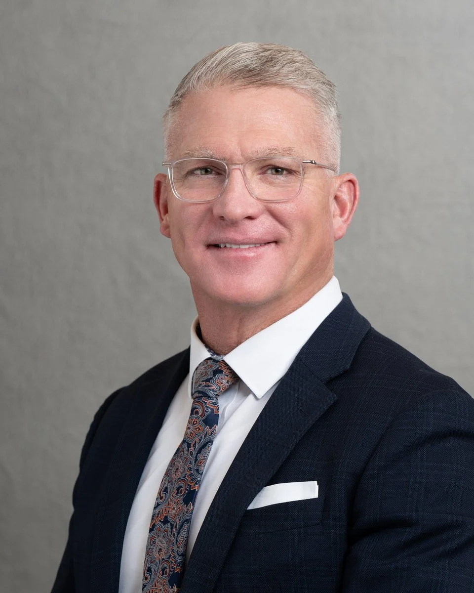 A professional headshot of a middle-aged man wearing glasses, a dark suit, a white shirt, and a patterned tie, smiling against a plain gray background.
