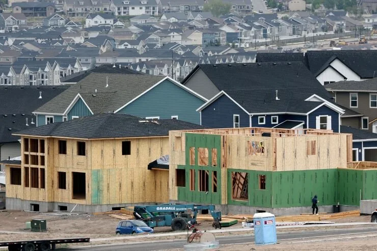 A new residential building under construction with wooden framing, set against existing completed houses in a neighborhood.