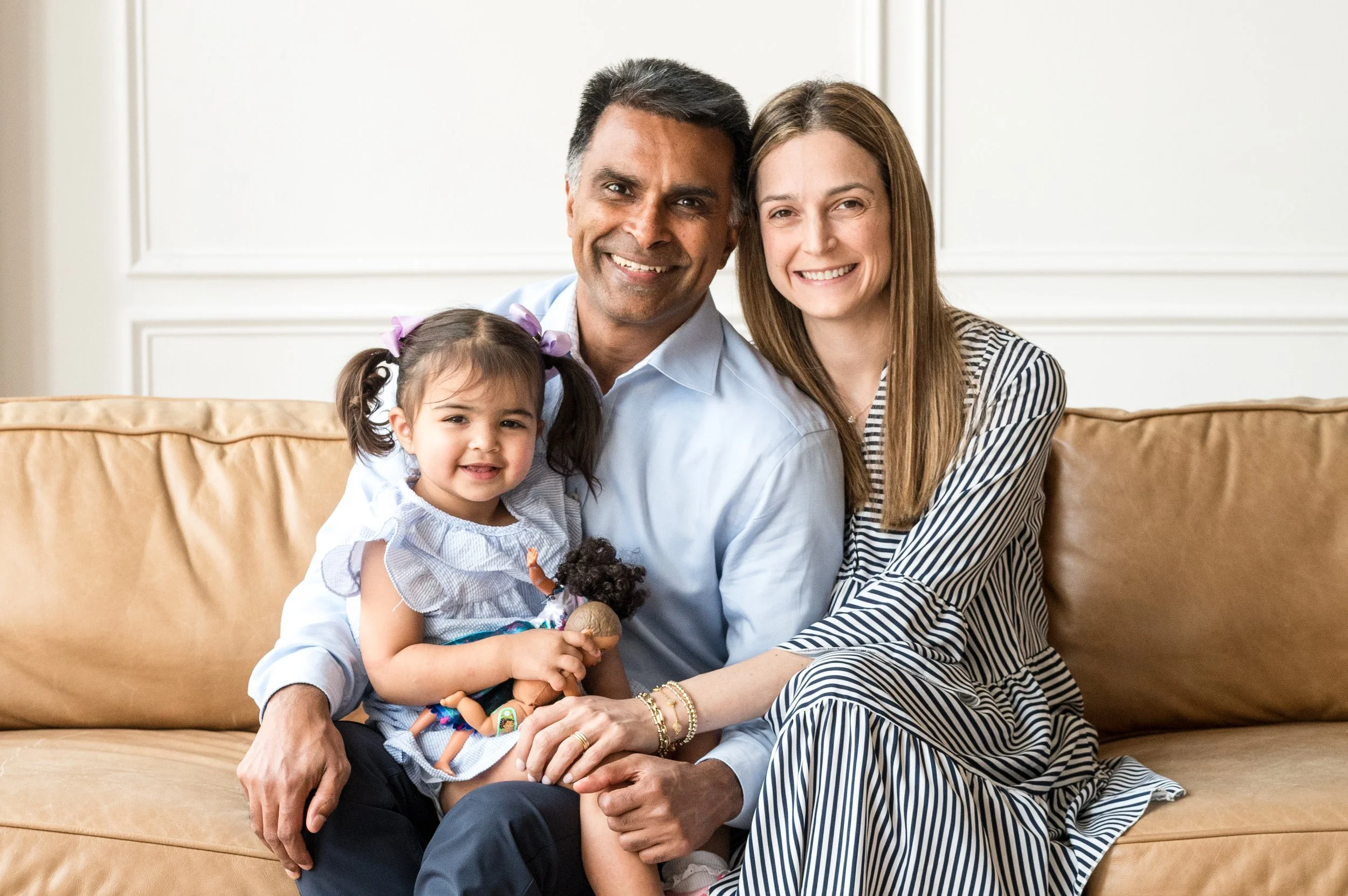 A multiracial family of four sitting on a beige sofa, smiling at the camera. The family includes a man, a woman, and two young girls, one of whom is holding a doll.