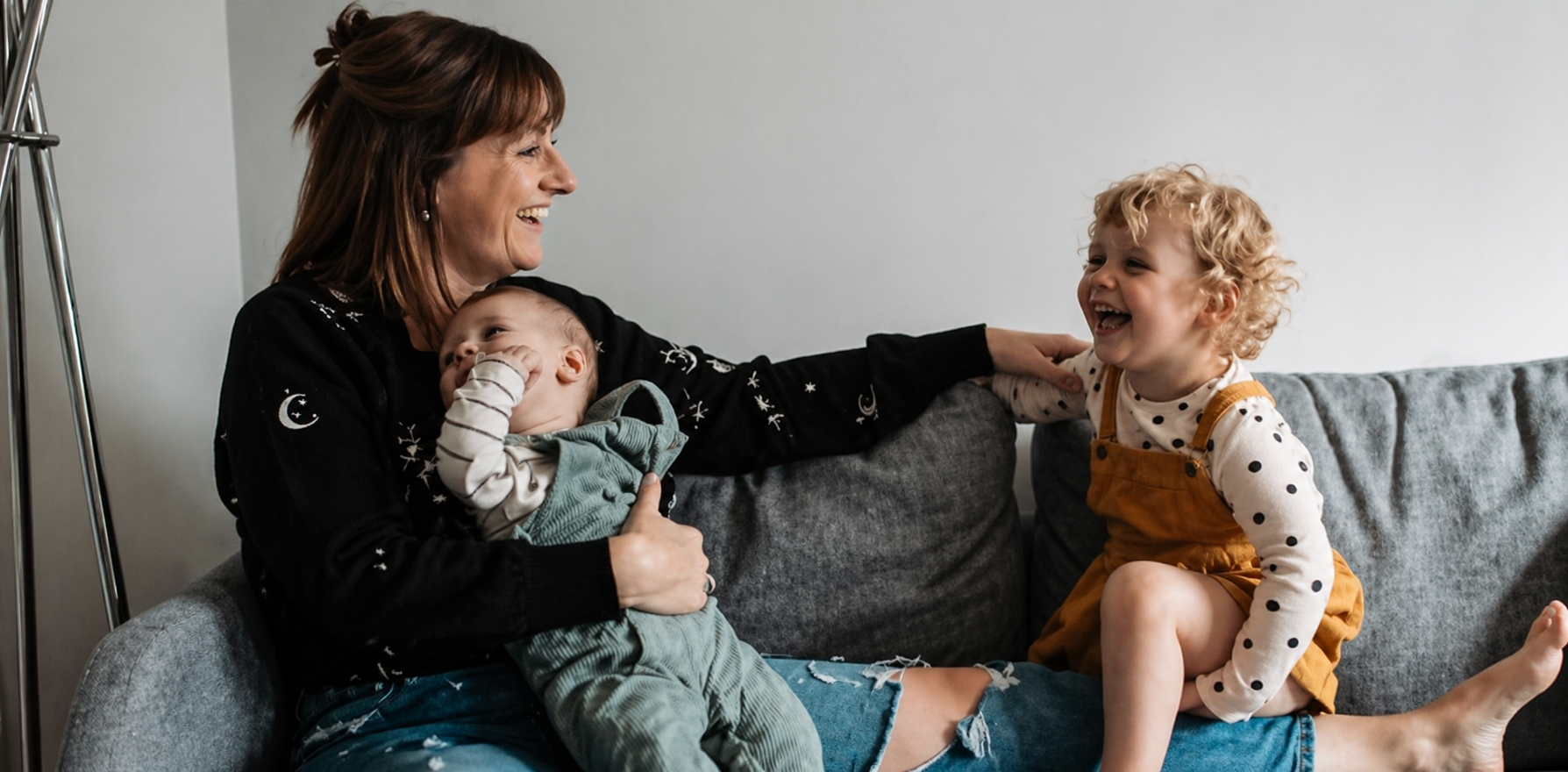 A woman with two children on a gray sofa, laughing and playing together. The woman has brown hair and is wearing a black sweater with moon and star motifs. The baby, held in her lap, is sucking on fingers, and the older child with curly blonde hair, wearing a polka-dotted shirt and mustard overalls, is smiling and touching the woman's arm.