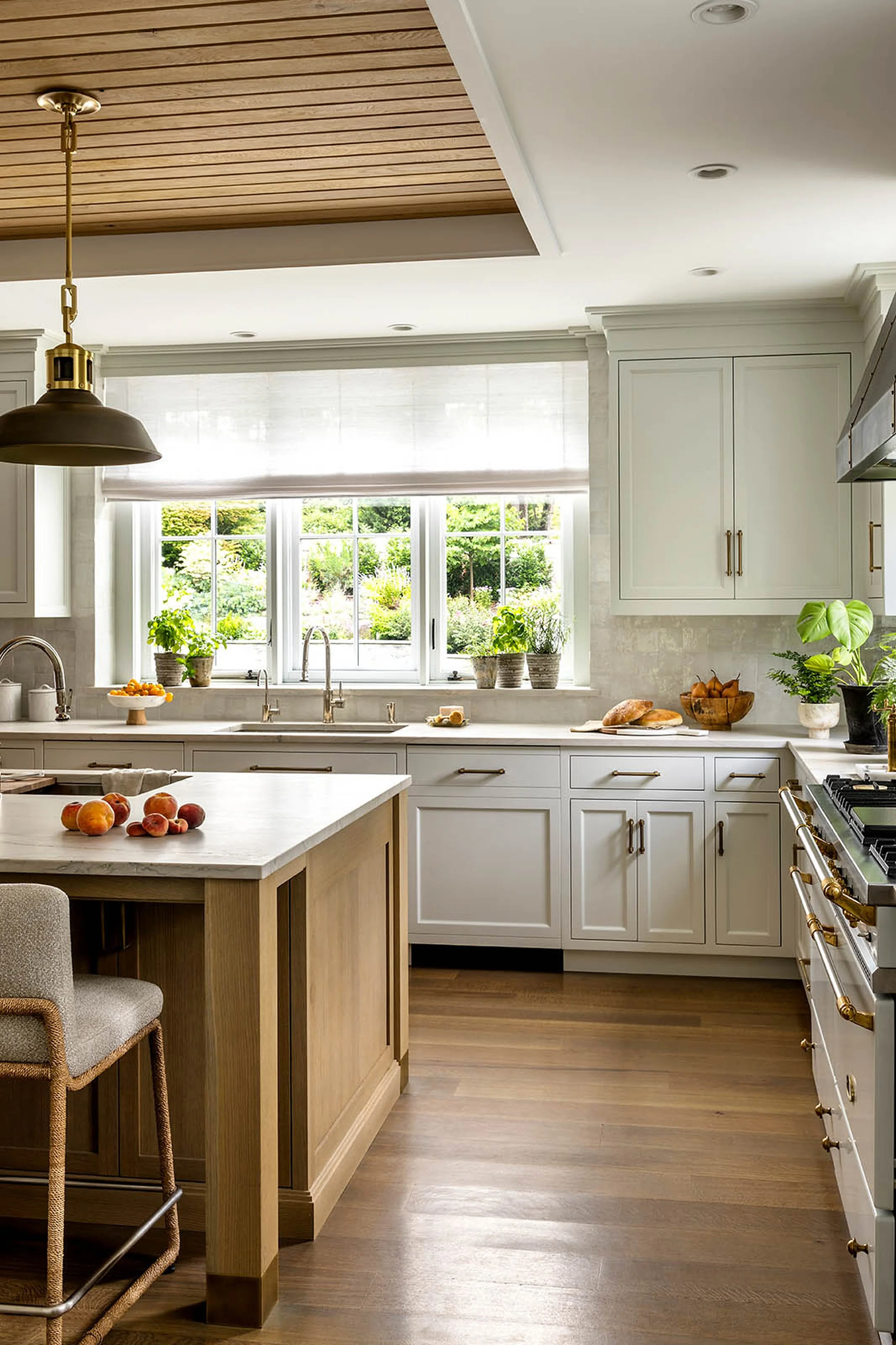 Kitchen with white cabinets, a large window, plants, a wooden island with peaches, a marble countertop, and a range with gold accents.