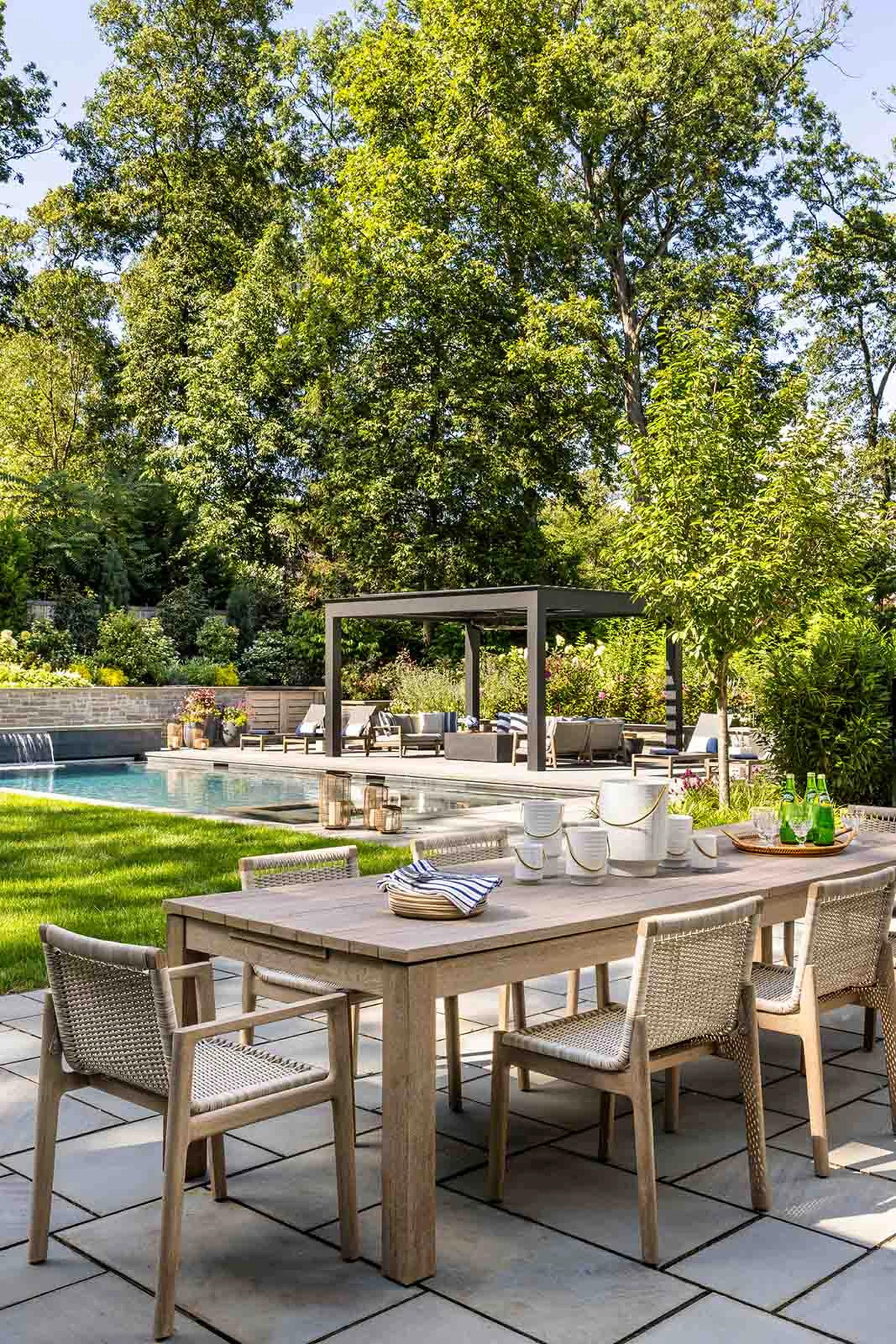 An outdoor patio with a large wooden dining table and chairs, overlooking a swimming pool surrounded by lush green trees and a garden.