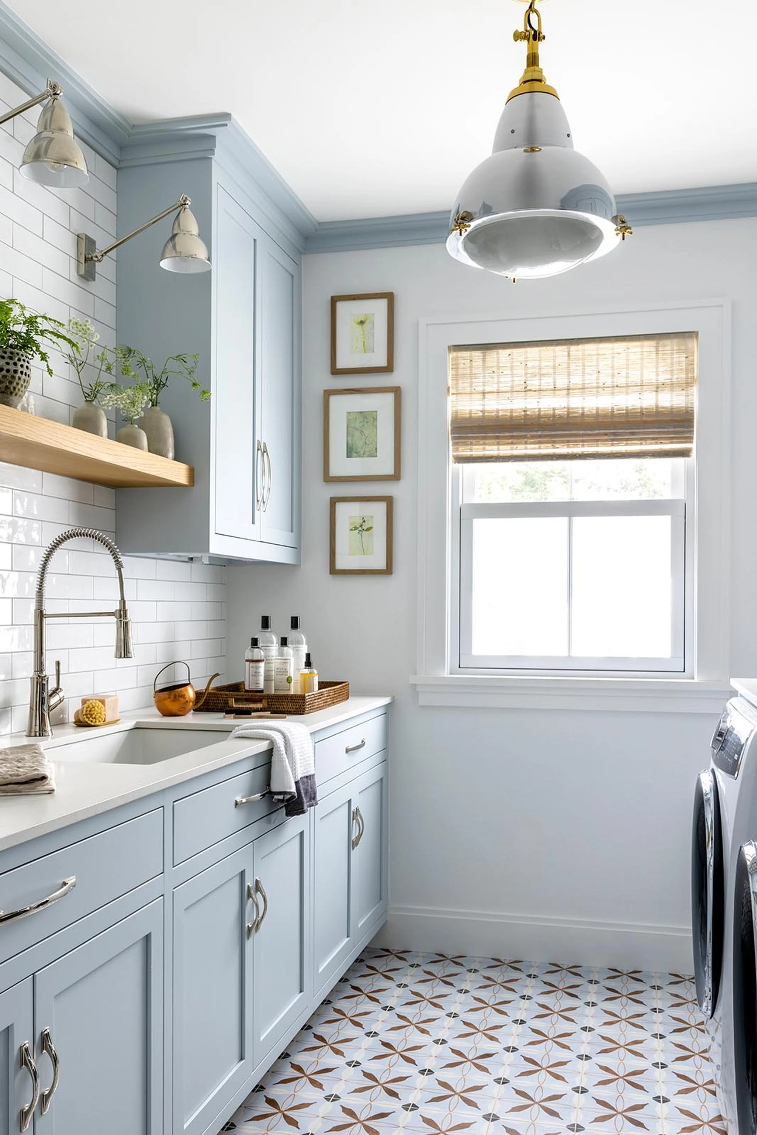 A bright, modern kitchen with white cabinets, a white tiled backsplash, and a window with a woven shade. Decor includes framed botanical prints and potted plants. A large metallic pendant light hangs from the ceiling.