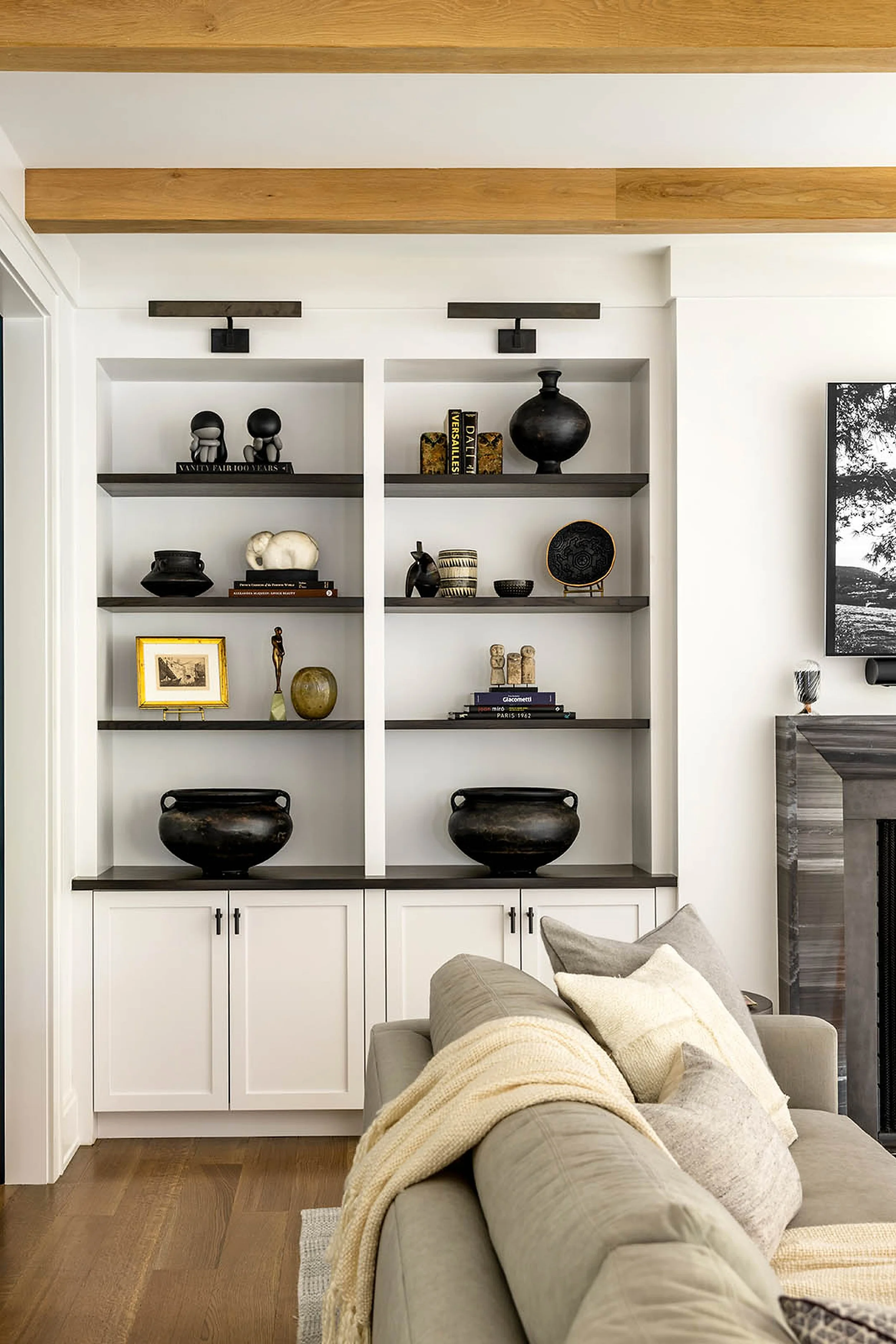 Living room with a built-in white bookshelf containing decorative objects and books, beige sofa with a throw blanket, wood flooring, and a partial view of a fireplace with a black and white framed photograph on the right.