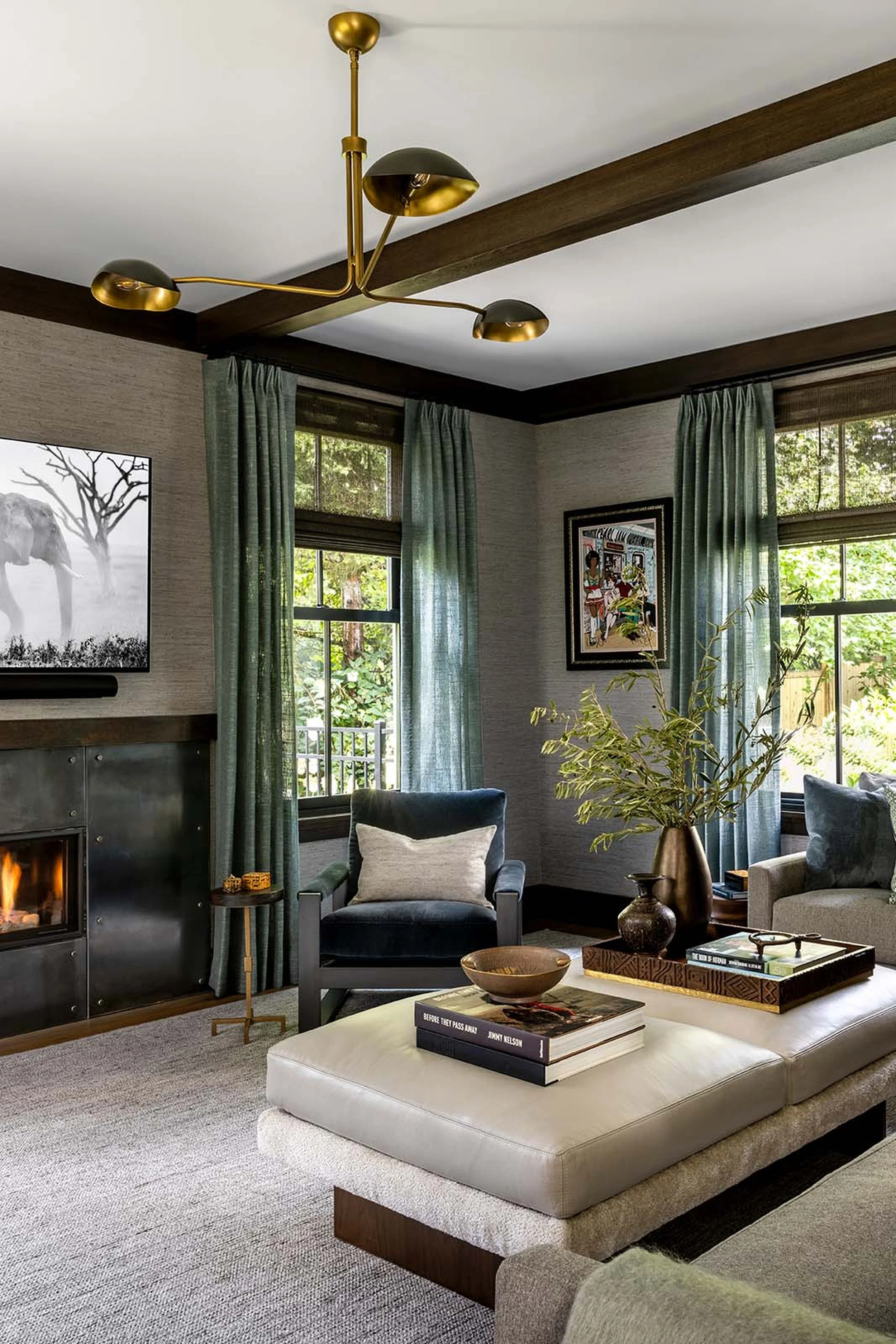 Living room with a modern gold chandelier, window with curtains, framed artwork, a fireplace, a gray armchair, a white upholstered ottoman with books and a decorative bowl, and a bronze vase with greenery on a side table.