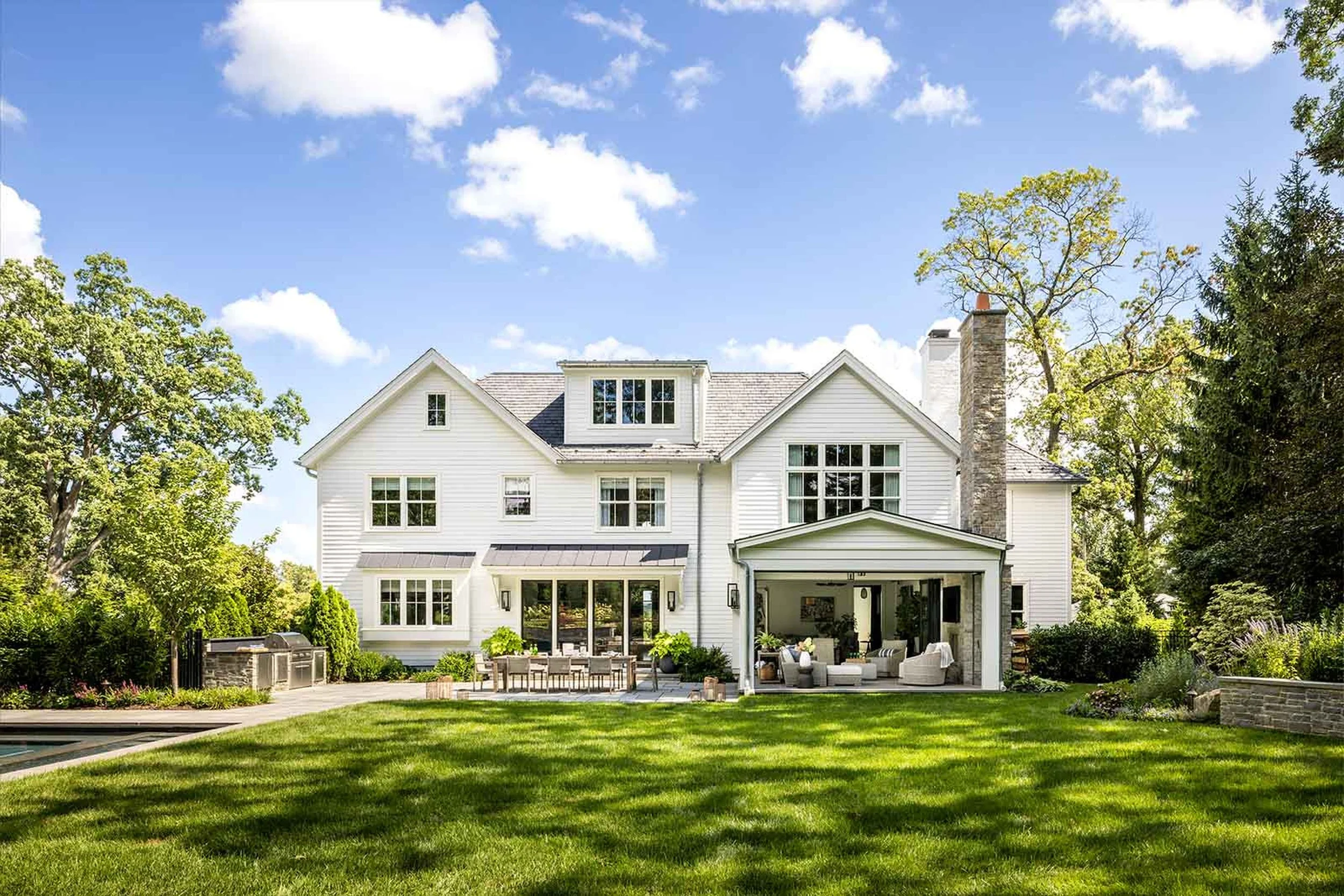 Large white house with multiple windows, a covered patio area with seating, surrounded by a lush green lawn and trees, under a blue sky with scattered clouds.