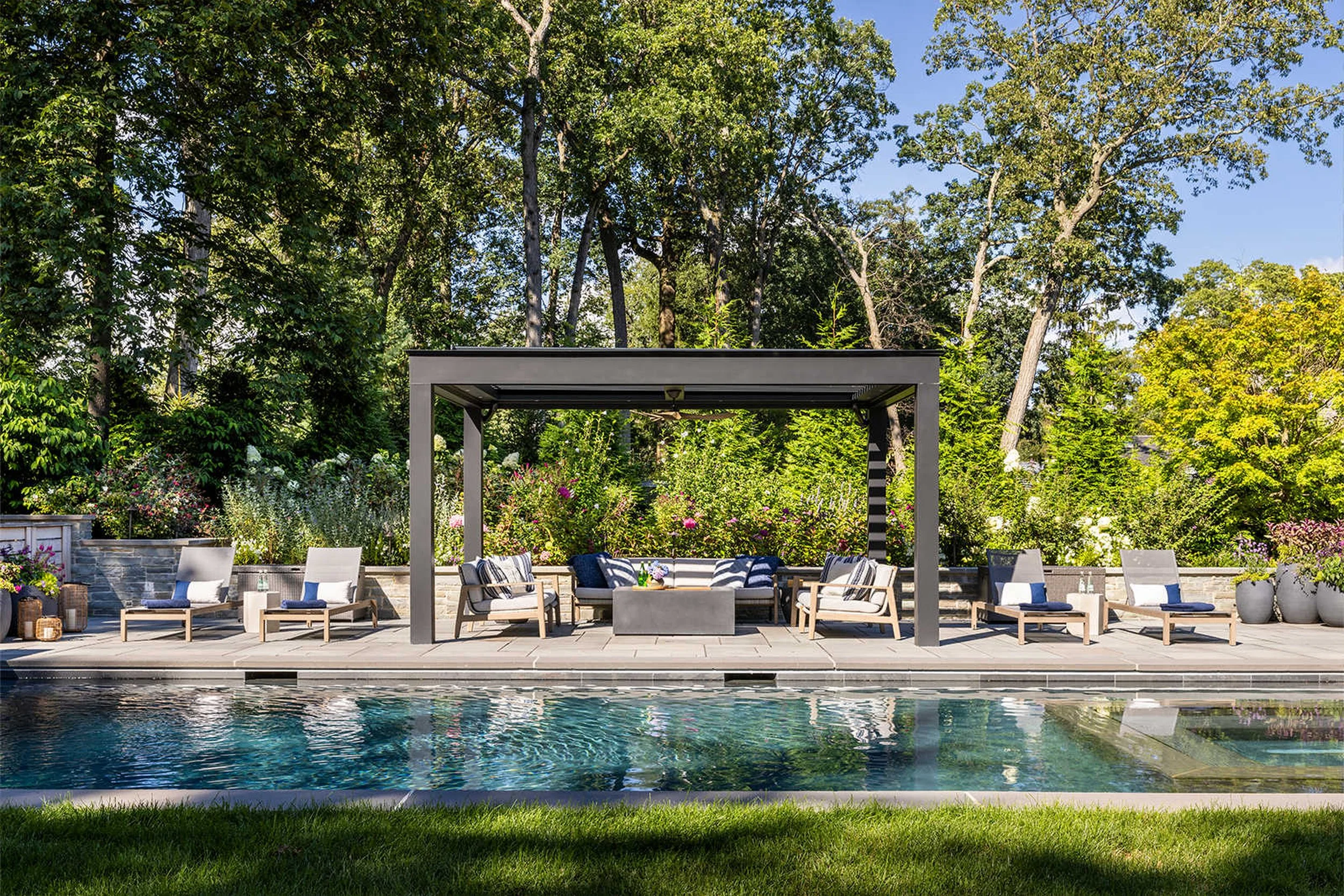 An outdoor living space featuring a modern pergola with seating, surrounding lush greenery, colorful flowering plants, and a swimming pool in the foreground under a clear blue sky.