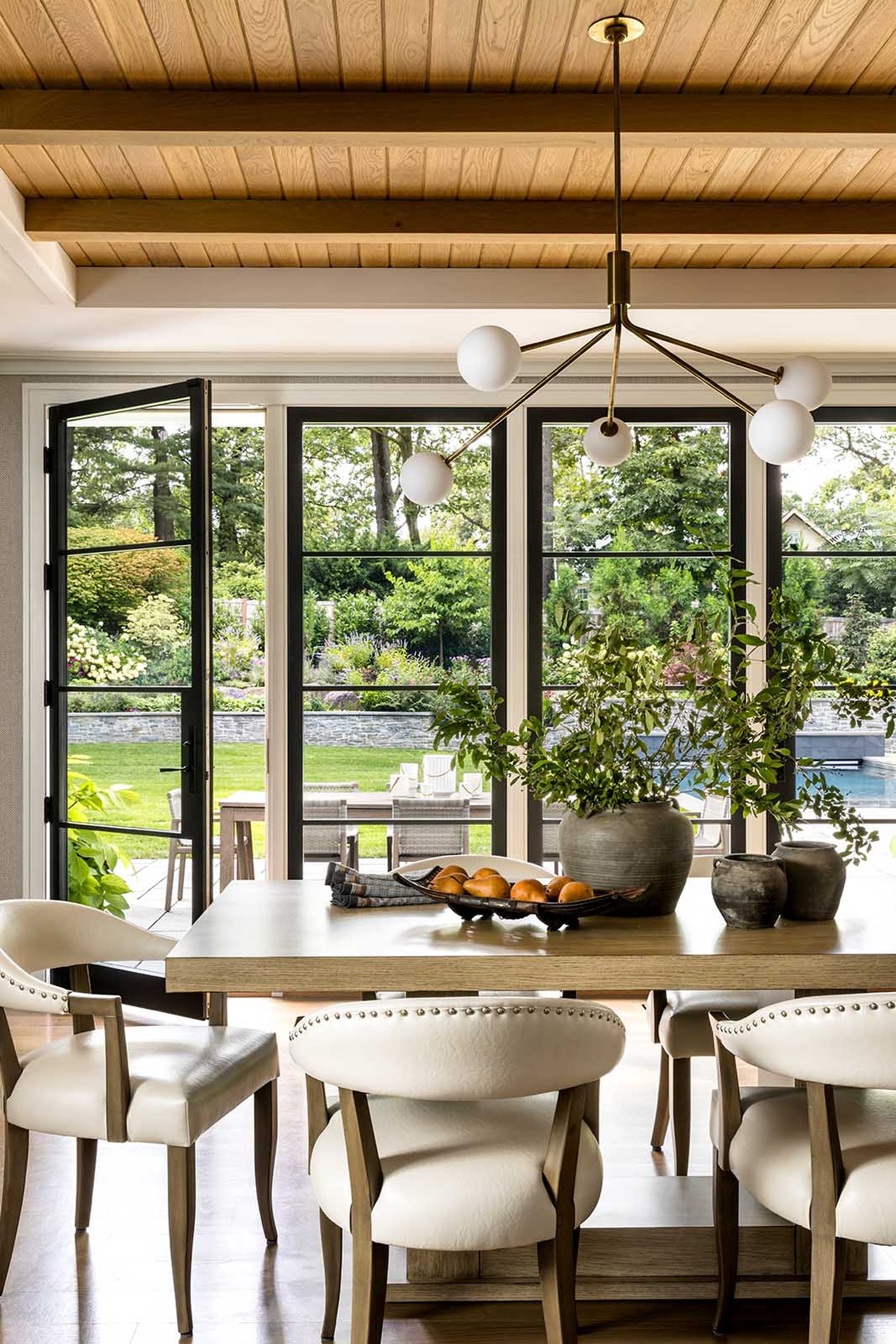A dining area with a wooden table and white chairs, decorated with a large potted plant, surrounded by large windows revealing a lush garden outside, and a modern pendant light fixture hanging from a wooden ceiling.