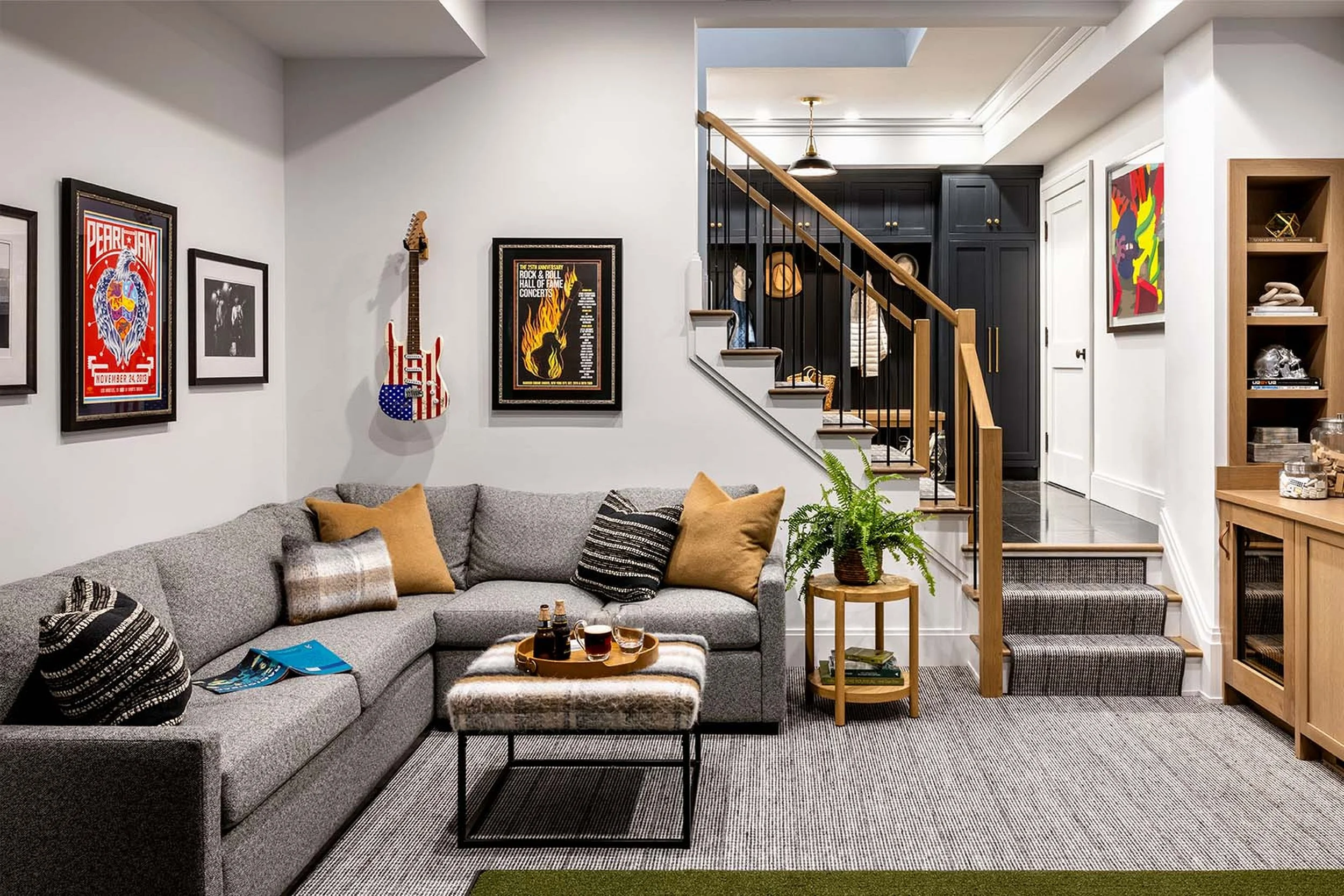 Living room with gray sectional sofa, black and white patterned pillows, a small wooden side table with plants, and wall art including posters and a guitar. Stairs with black balustrade lead to an upper level, and dark cabinetry is visible in the bac
