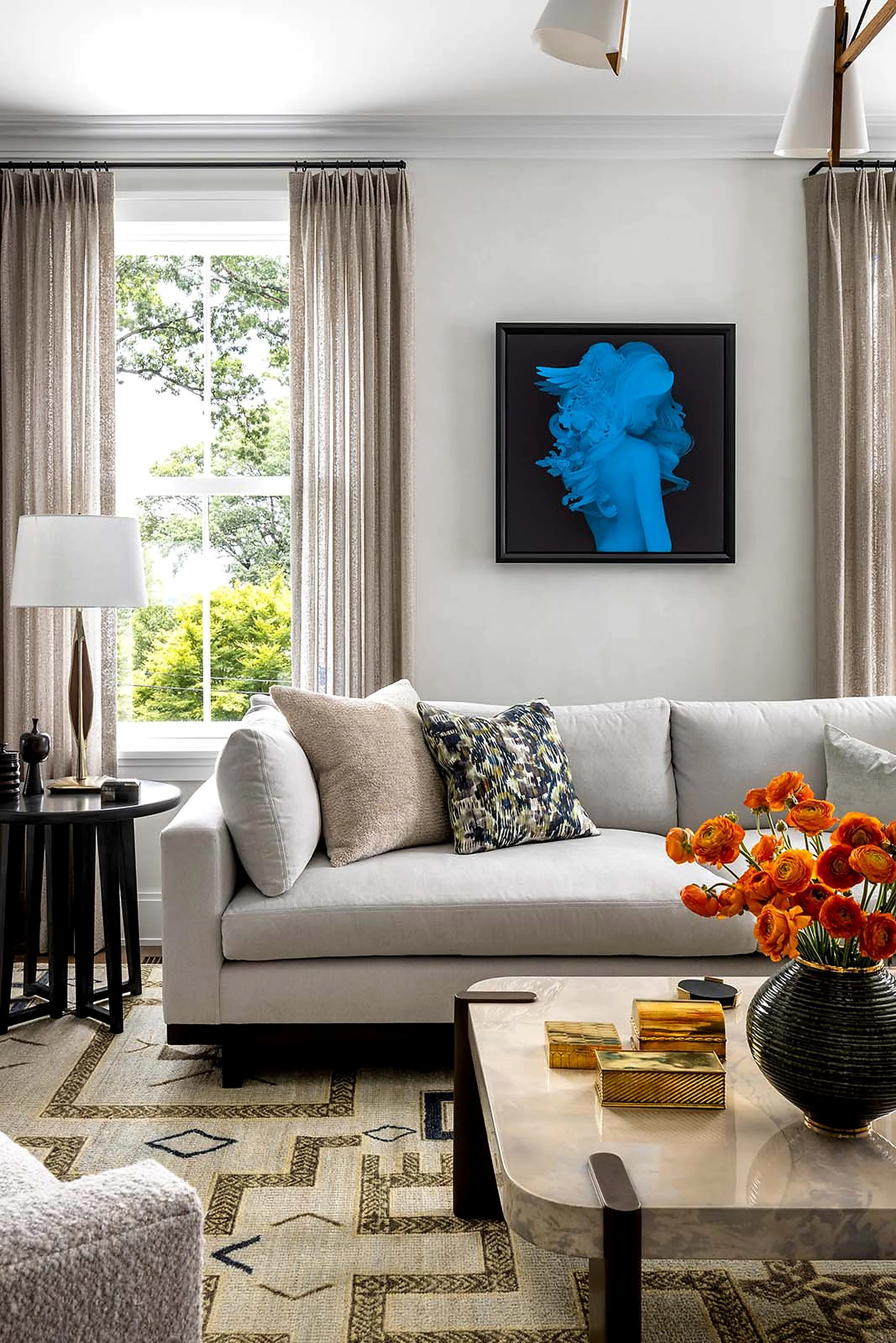 Living room with a white sofa, decorative pillows, a marble coffee table with gold accents, an orange flower arrangement, a black framed artwork of a woman with blue hair, beige curtains, and a large window with a view of green trees.