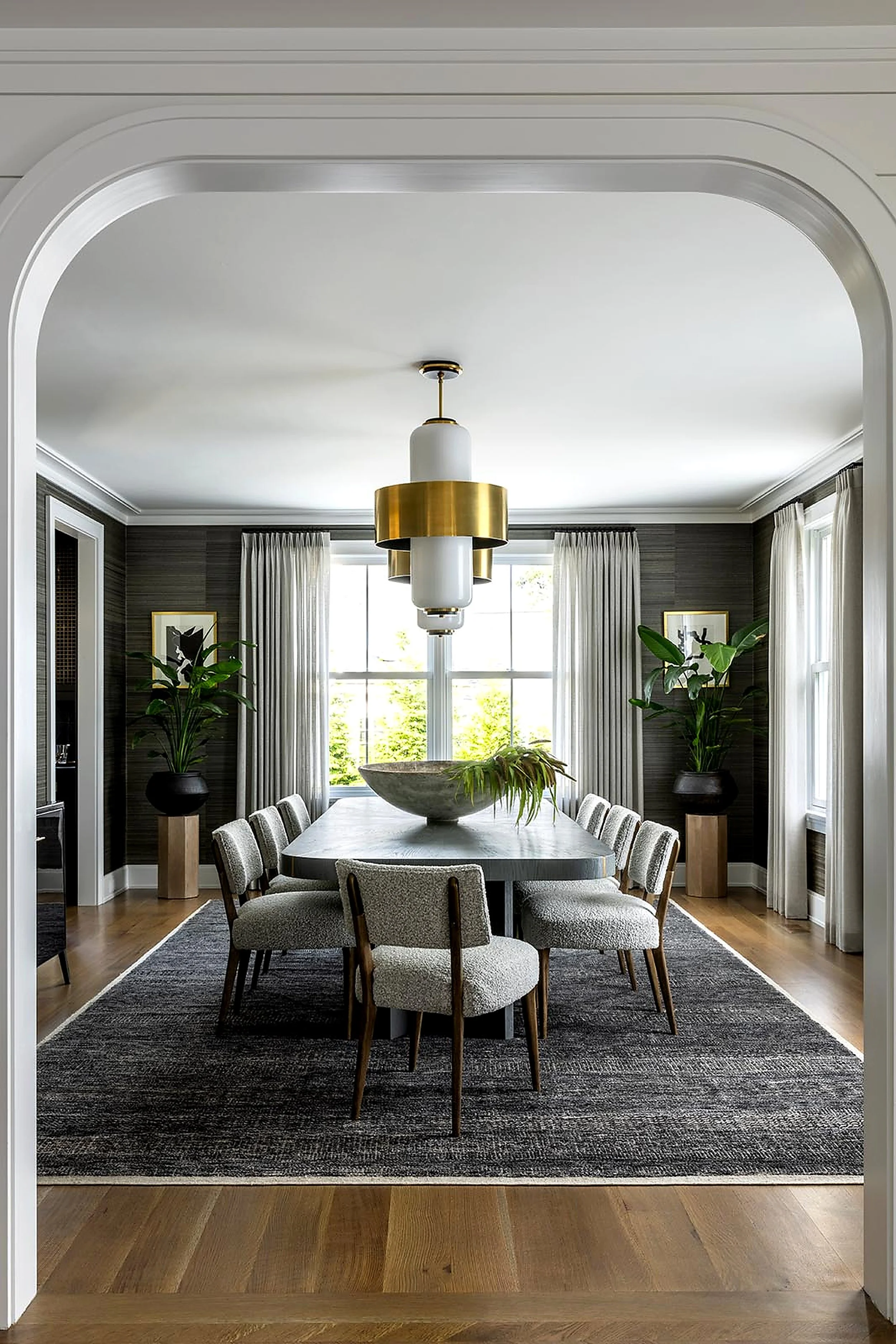 This is a modern dining room with a grey color palette. It features a large rectangular table with eight upholstered chairs, a black and grey area rug, large potted plants, and wall art. A large window with curtains allows natural light to fill the room, and a contemporary chandelier hangs above the table.