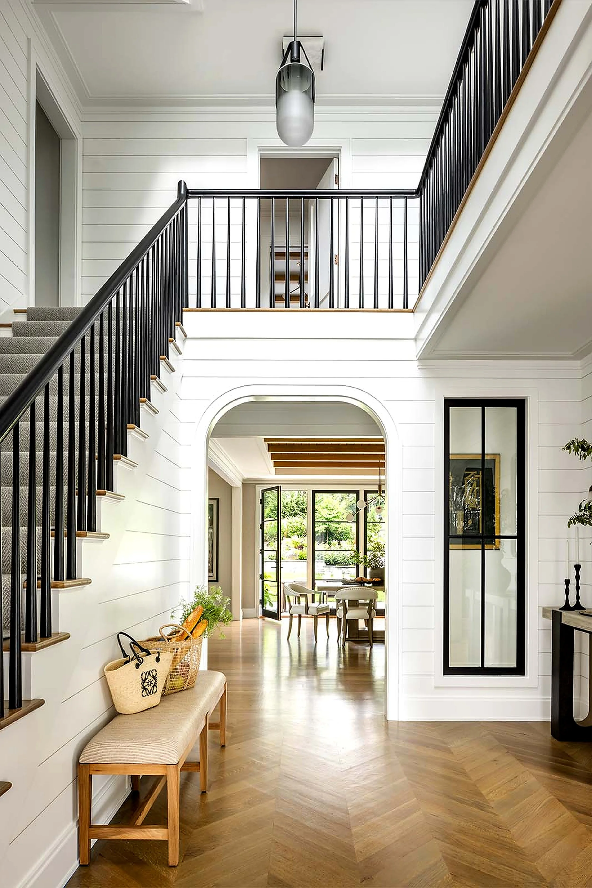 Interior view of a modern home foyer with wooden flooring, white paneled walls, a staircase with black railing, and a sitting area with a beige bench and baskets. Visible dining area and living room through an arched doorway.