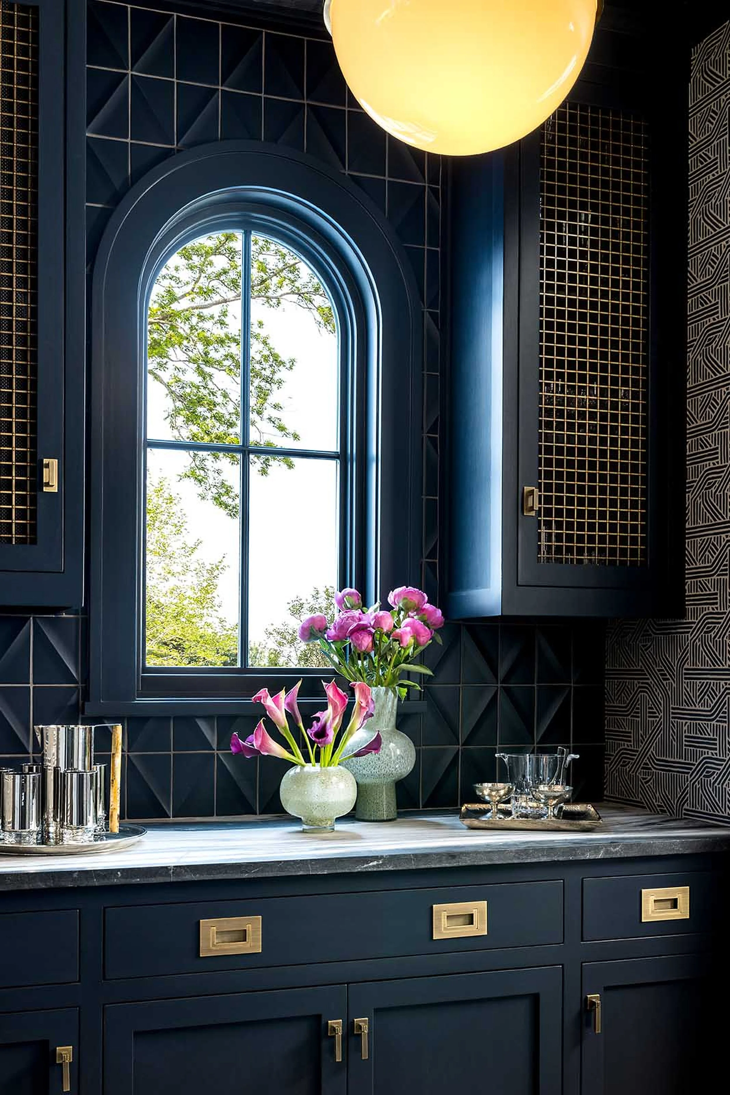 A kitchen with dark blue cabinets and gold hardware, a marble countertop, a window with an arched top showing green trees outside, decorative gold grilles on the walls, a vase of pink flowers on the counter, and various glassware and metal containers