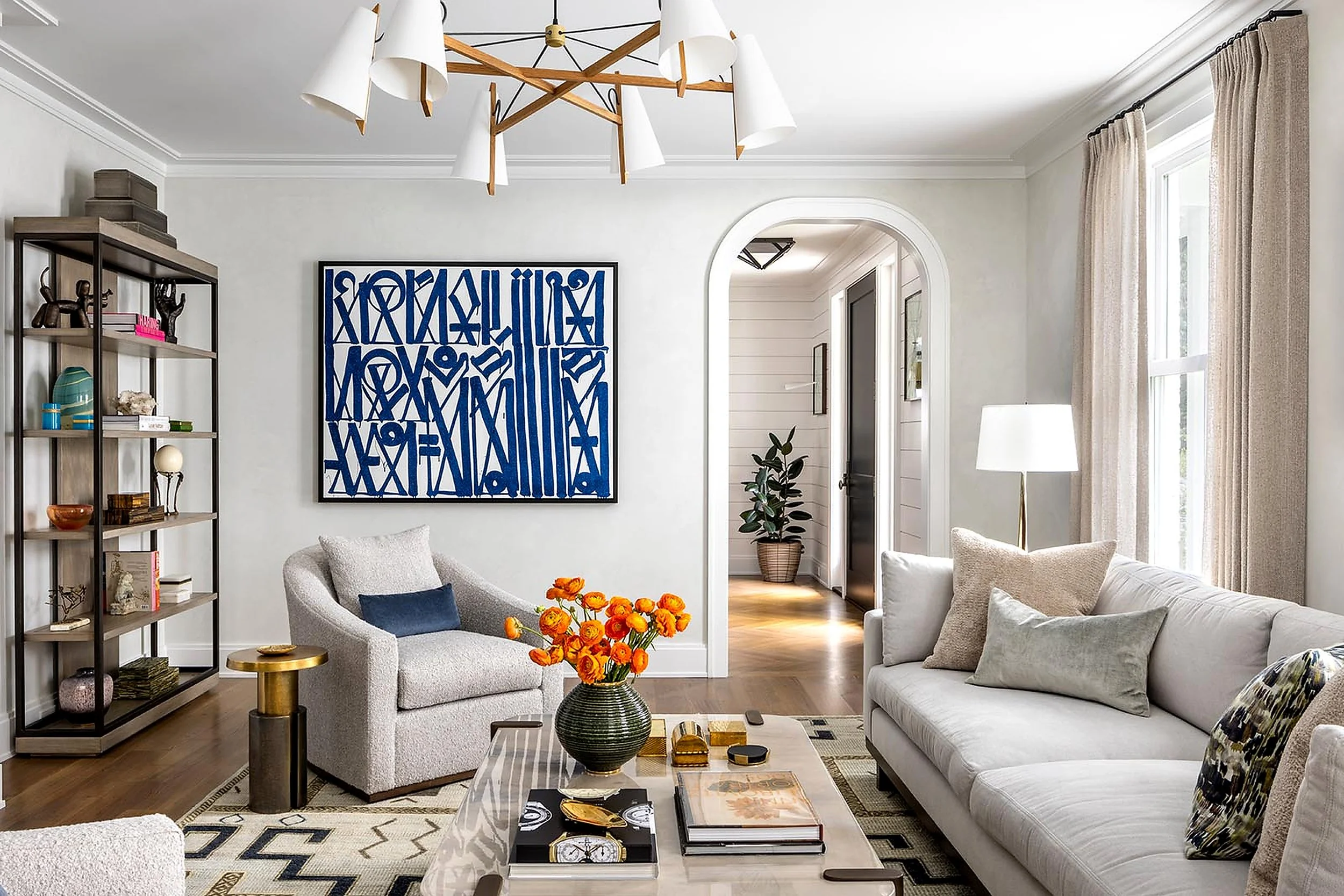 A living room with large windows, beige curtains, a light-colored sofa, a white armchair with a blue pillow, a black and gold coffee table with books, a vase of orange flowers, a patterned rug, a black floor lamp, a wooden bookshelf with decorative i