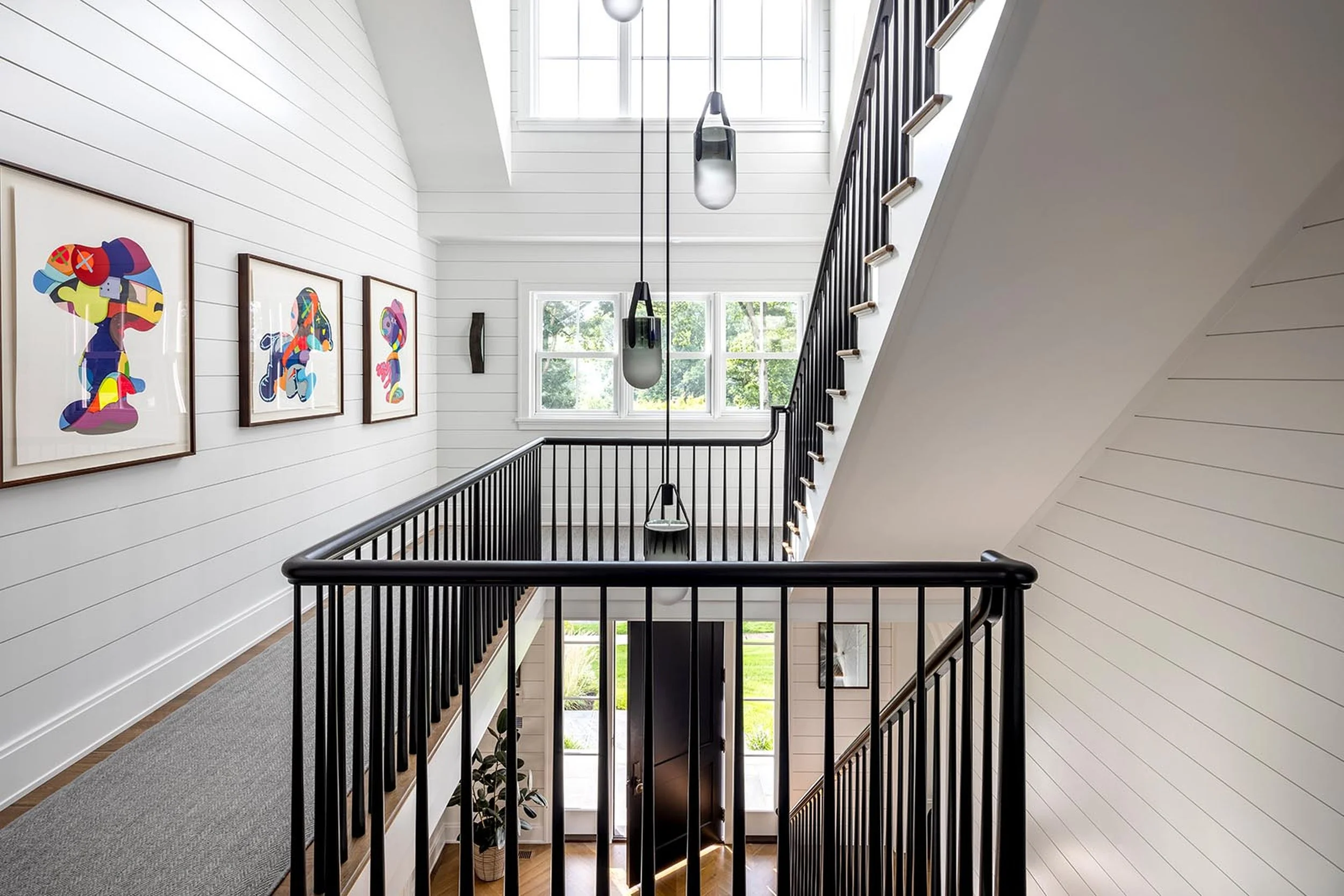 A staircase landing in a modern home with white shiplap walls, large windows, and black railing. Artworks with colorful abstract designs hang on the wall. Hanging pendant lights illuminate the area.