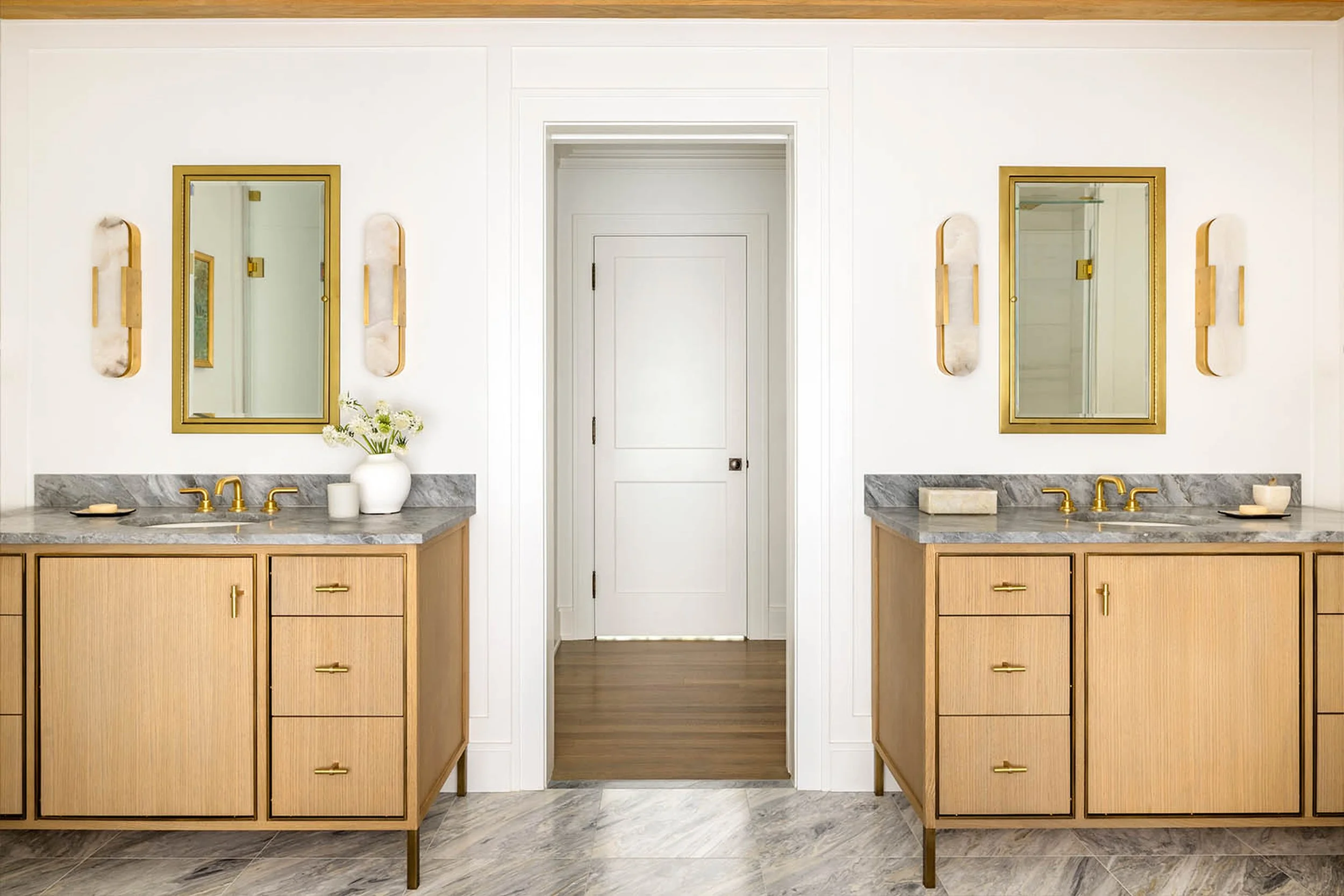 A bathroom vanity with two sinks, mirrors with gold frames, and wooden cabinetry with gold handles. A vase with white flowers is on the left side on the countertop, with soap and other small items nearby. Two wall-mounted light fixtures are beside th