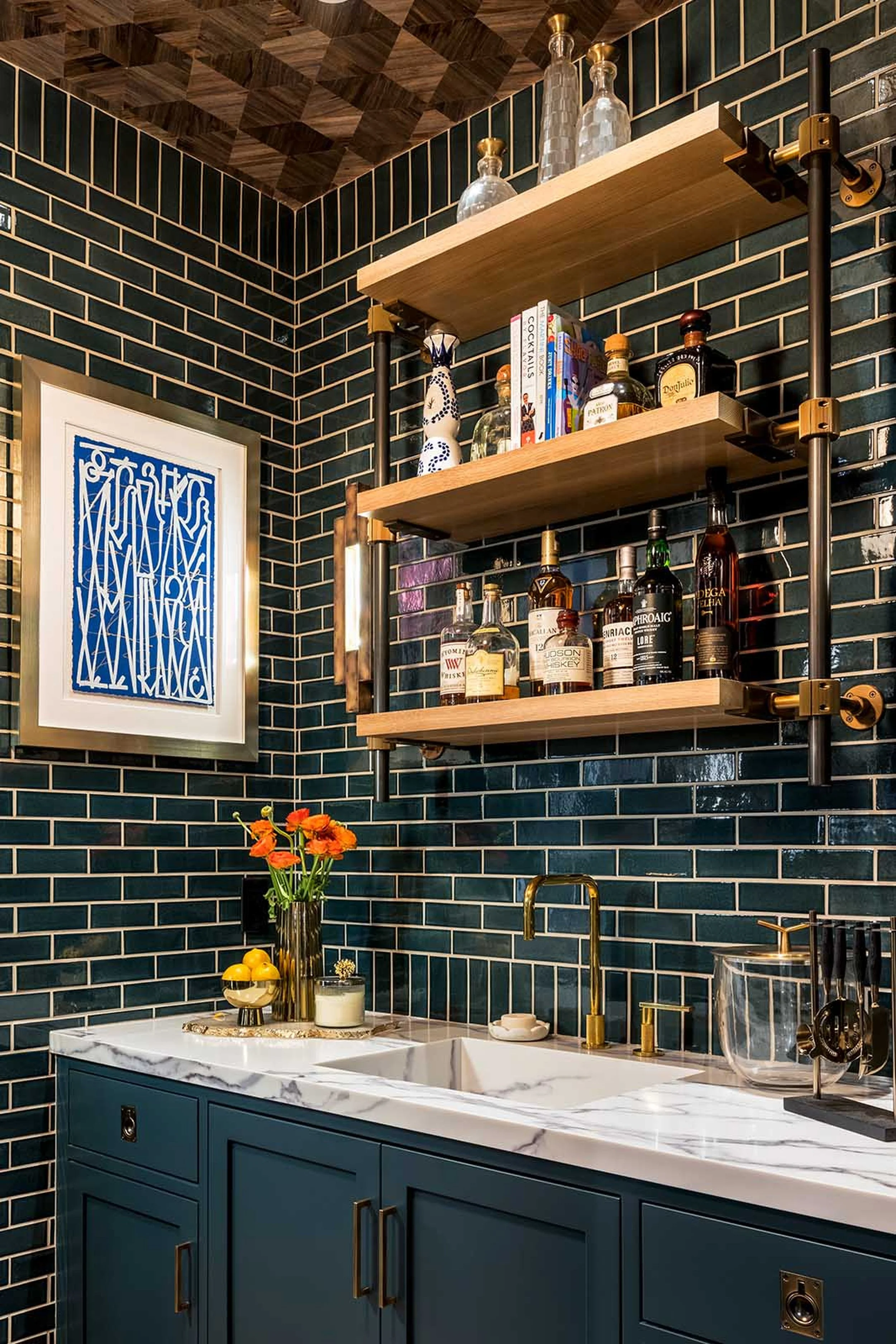 A modern kitchen with dark blue cabinets, a white marble countertop, and black subway tile walls. There are wood and brass open shelves holding bottles and books, with a picture frame on the wall and an orange flower arrangement on the countertop.